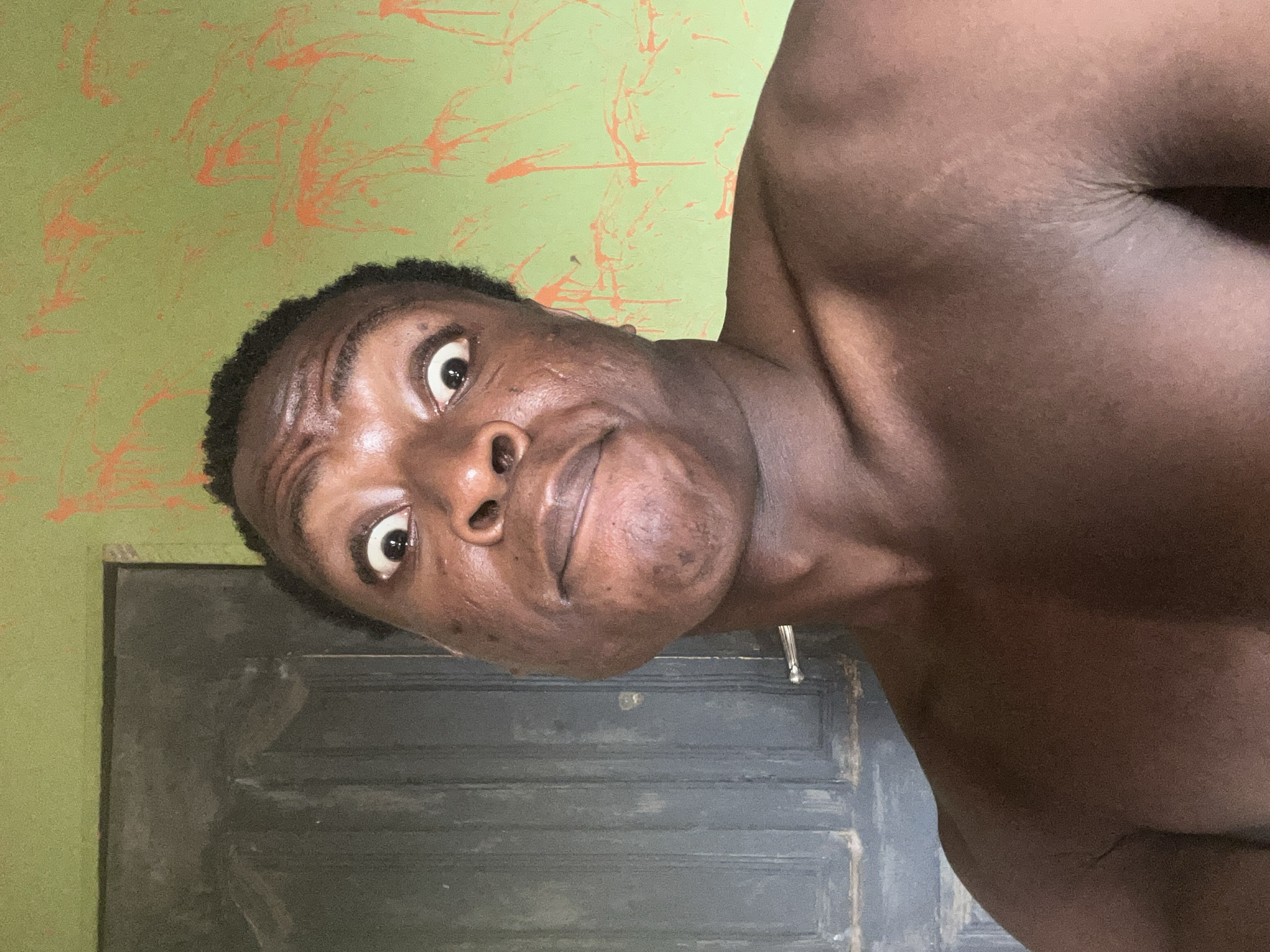 A close-up, rotated image depicts a dark-skinned person with wide eyes and a slightly puckered expression. The person appears to be shirtless. The background features a green wall with orange splatter-like patterns. Behind the person, there's a gray door with horizontal grooves. The location is Unknown, Unknown.