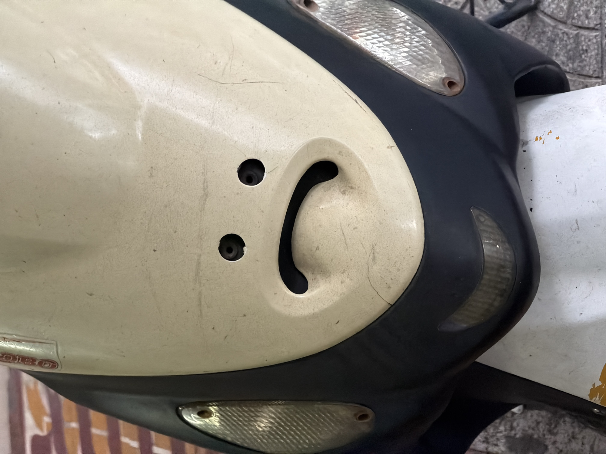 The image shows a close-up of the front of a motorcycle. The motorcycle has a cream-colored front panel with a black trim. On the front panel, there are two small holes and a larger, curved opening. Above the panel, there is a clear headlight and a black section. There is also a white section visible on the right side. The background is a blurry mix of tiled surfaces and other objects. Given the prevalence of motorbikes in Đà Nẵng, Vietnam, and the general style of the motorbike, it is possible the photo was taken there, but this cannot be stated with certainty.