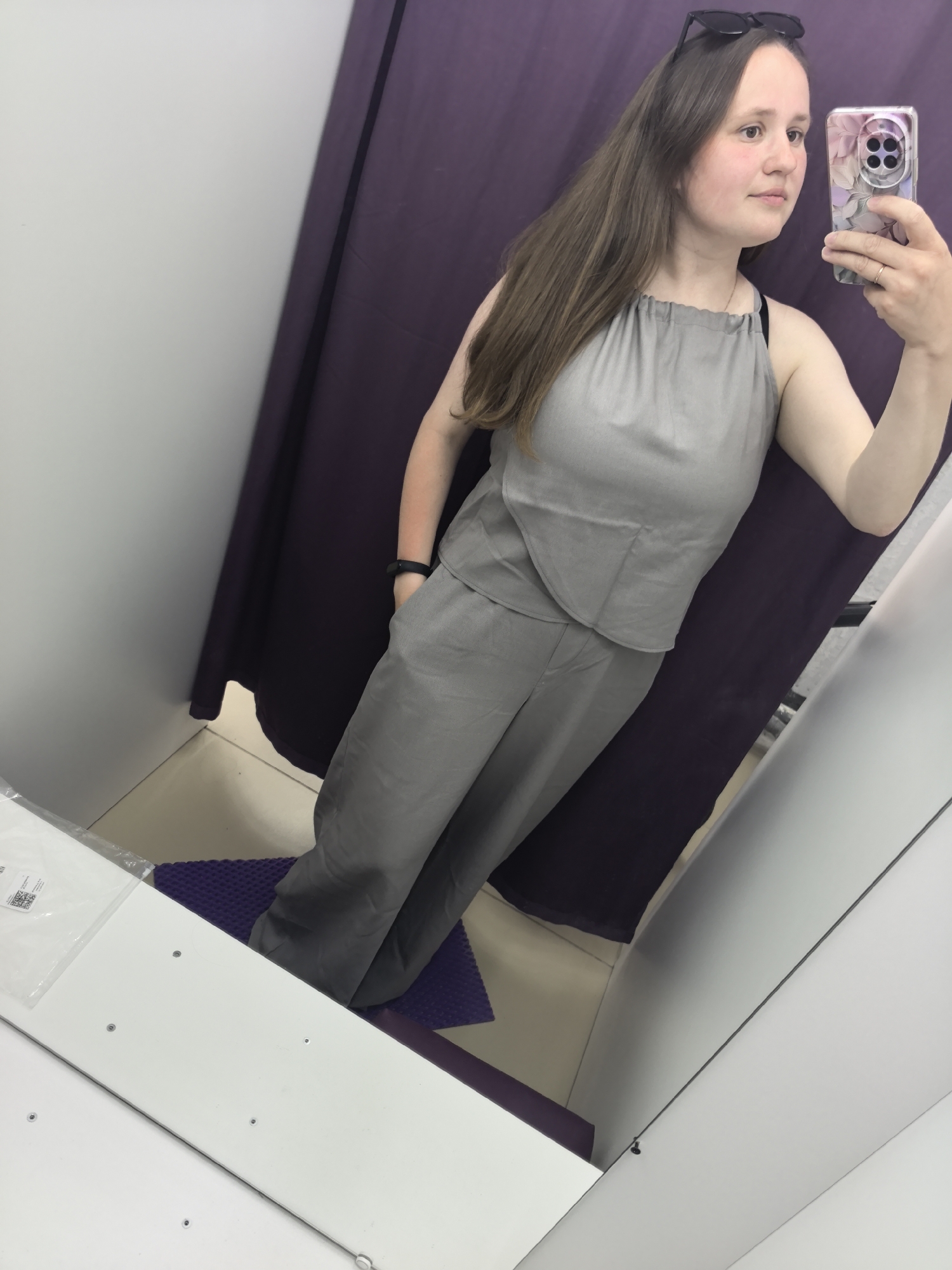 A young woman is taking a selfie in what appears to be a dressing room. She is wearing a gray, sleeveless two-piece outfit. Her hair is long and brown, and she has sunglasses on her head. She is holding a smartphone with a patterned case in her right hand. The background consists of a white wall and a purple curtain. A purple mat is on the floor. The location is Фролы, Russia, according to additional information.