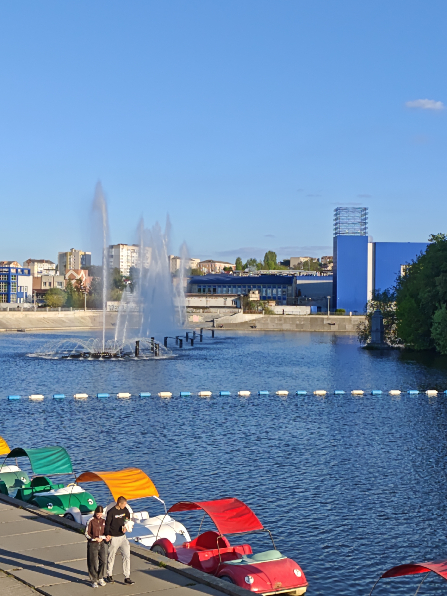 The image shows a waterfront scene in Vinnytsia, Ukraine. A body of water, likely the Southern Buh river, features a fountain in the middle. A line of colorful paddle boats, resembling small cars, are docked along the concrete bank. A couple, a man and a woman, are walking along the promenade next to the boats. The man wears a dark sweatshirt and light-colored pants. The woman is in a track suit. Buildings are visible in the background, including a tall blue structure with a metal framework on top. The sky is clear and blue.