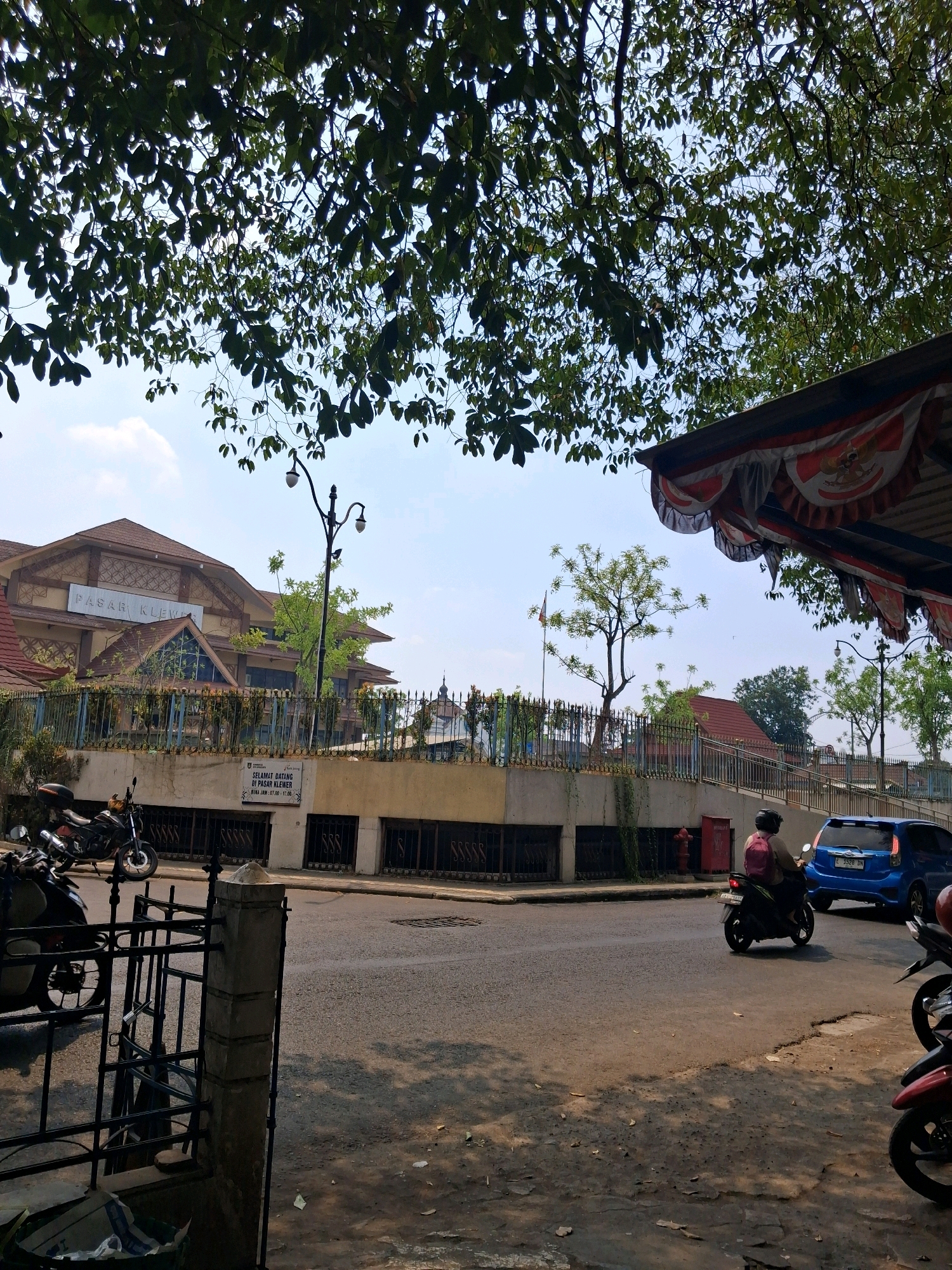The image shows a street scene in Surakarta, Indonesia, likely near Pasar Klewer market. In the background, a large building with a brown roof is visible, bearing the sign "PASAR KLEWER". A fence with metal railings separates the street from the market building. Several motorcycles are parked on the left side of the street, and one is in motion on the right side, carrying a person wearing a pink backpack. A blue car is also visible on the right, driving away from the viewer. A fire hydrant is mounted on the wall of the building. The foreground is dominated by trees with dark green leaves, casting shadows on the street. The sky is clear with some clouds. The overall impression is that of a typical day in a bustling Indonesian market area.
