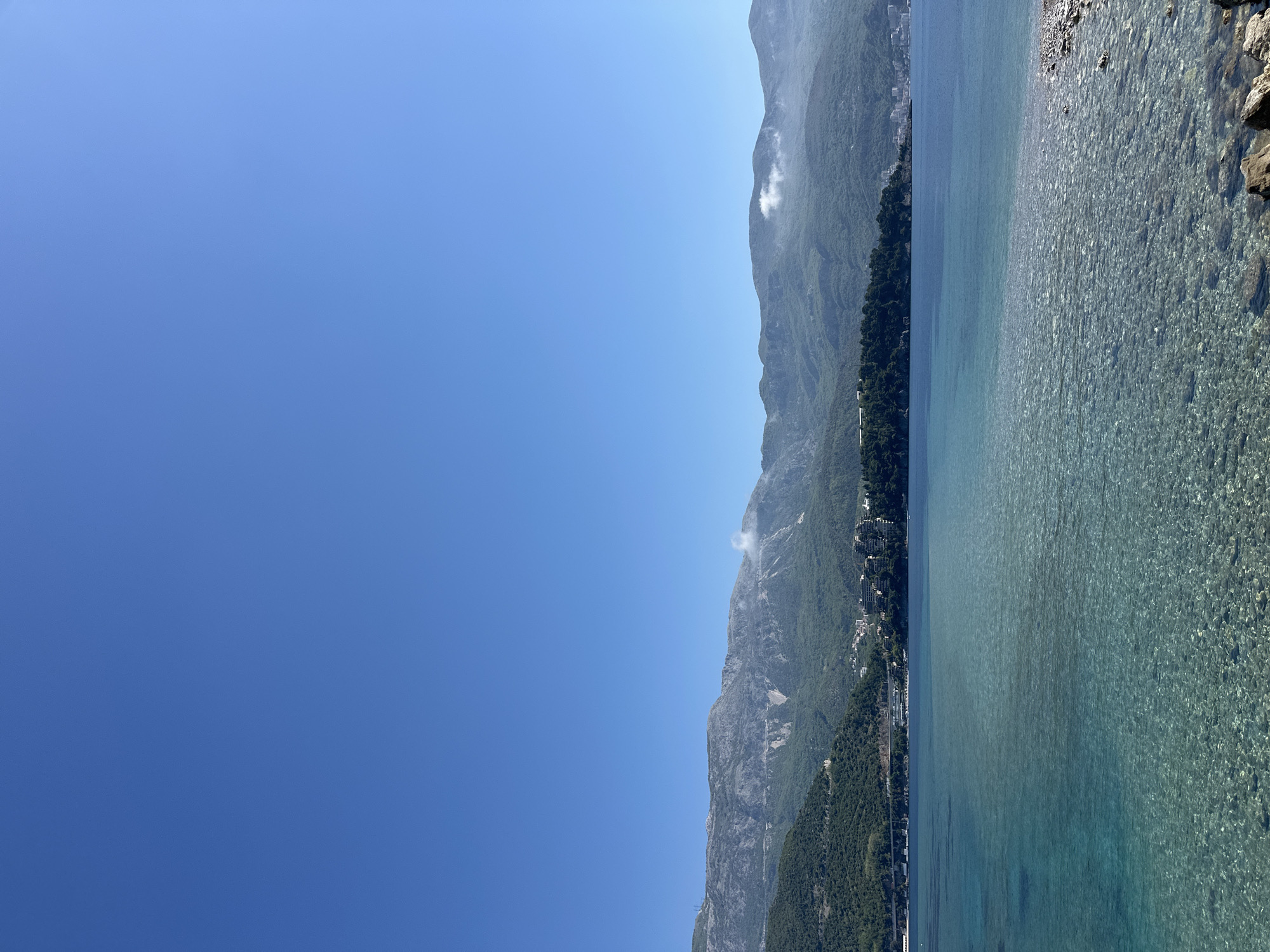 The image shows a view of the Adriatic Sea along the coast of Budva, Montenegro. High, forested mountains rise sharply along the coastline. A small town with closely packed buildings lies at the base of the mountains along a narrow strip of land. The water is clear and turquoise, with visible rocks near the shoreline. The sky is clear and blue.