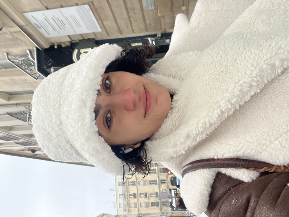 The image displays a close-up portrait of a female individual captured outdoors in an urban setting during snowfall. The subject has dark, curly hair, brown eyes, and is looking directly forward with a subtle, closed-mouth smile. Her cheeks exhibit a slight redness, consistent with cold weather exposure. She is dressed in winter attire, including a white, fluffy, sherpa-style bucket hat and a matching white, plush scarf or collar, which appears to be part of a coat. A portion of a brown, possibly leather or shearling, jacket is visible on her right shoulder, featuring a white sherpa lining. A white wireless earbud is present in her left ear, alongside a gold-toned hoop earring. A brown braided strap, likely from a bag, is visible over her right shoulder.
The background features classic Haussmannian-style architecture, identifying the location as Paris, France. Multi-story buildings constructed of light-colored stone with dark window frames and ornate iron balconies are visible. Snow dusts the balconies and ledges, and faint snowflakes are discernible in the ambient air under an overcast sky, indicating cold, wintry conditions. On the right side of the image, a vertical white sign with text, partially obscured, shows "RVP" at its upper section and additional French script below, suggesting an official notice. Beneath this, a dark sign with a white "21" is partially visible. In the distant left background, blurred figures of people and the facades of other buildings suggest a populated street.