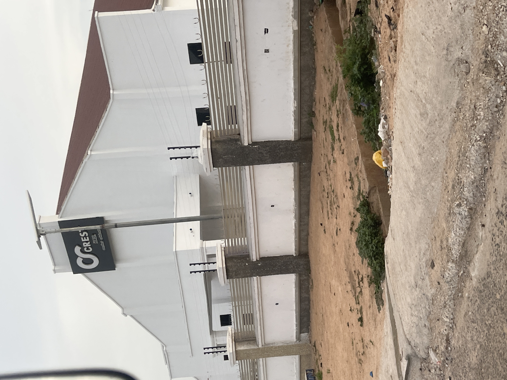 This image, set in Howayi, Nigeria, depicts a modern, multi-story building with a clean white facade and a contrasting brown sloped roof. The structure features multiple levels, each with railings or balconies made of vertical light-colored bars. Several black spikes, likely parts of an electric fence, are visible on top of the walls and pillars, suggesting security measures.
A prominent black rectangular sign is mounted on a pole extending from the building. It clearly displays the text "CREST HALL" in large white lettering, with "CREST" being more stylized. Below this, the words "comfort & relaxation" are inscribed in a smaller, cursive-like font.
To the right of the building, the scene transitions to an unpaved, uneven dirt road or ground. This area is characterized by packed earth, small rocks, and sandy patches. Sparse green vegetation, consisting of bushes and weeds, grows along the edges of this dirt area, particularly in a slight depression. Some litter, including a distinct yellow plastic object, is scattered on the ground.
The sky is overcast and bright, indicating daytime conditions without direct sunlight. There are no people visible, and no specific activity or event is taking place, presenting a static view of the building and its immediate, somewhat undeveloped surroundings.