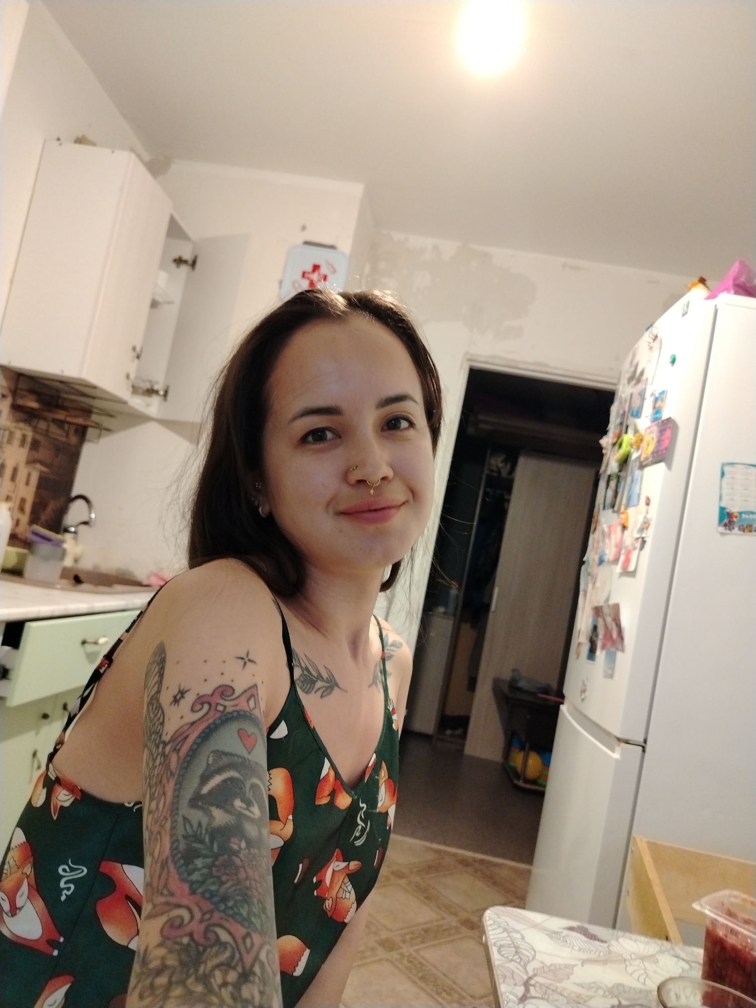 A young woman with dark hair and a nose ring is posing for a photo in a kitchen. She is wearing a green tank top with a fox pattern. Her left arm has a large tattoo of a raccoon in a frame, and smaller tattoos are visible on her chest and shoulder. The kitchen has white cabinets, a sink, and a white refrigerator covered with magnets and papers. A first-aid kit is mounted on the wall above the refrigerator. A doorway in the background leads to another room. The location appears to be in Noviy, Russia, based on the "Новый" that is possibly seen on the refrigerator.