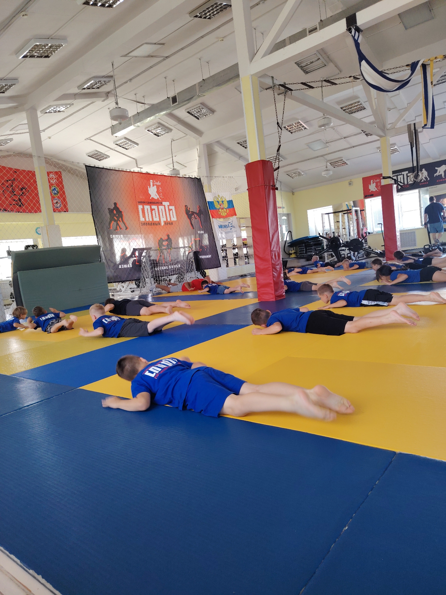 The image depicts an indoor martial arts or gymnastics training facility, likely in Chaikovsky, Russia, as suggested by the flags visible. The room is spacious with a high ceiling featuring fluorescent lighting and ventilation systems. The floor is covered with blue and yellow mats, arranged in a grid pattern.
A group of young individuals, predominantly boys dressed in blue sports uniforms with white lettering, are engaged in physical exercises. Most of them are lying prone on the mats with their faces down, performing some form of core or strength training. Their body positions suggest they are holding a plank or a similar exercise. Some are seen with their legs extended, while others have their arms positioned under their chins.
In the background, there are larger mats stacked against a wall, and a wrestling net is set up. Several banners and flags are displayed on the walls, some bearing Cyrillic text and what appear to be national or club insignias, including the Russian flag. Exercise equipment, such as weight machines and other gym apparatus, are visible in the further background, indicating a well-equipped training center. A man in a dark blue shirt and shorts is standing near the entrance on the right side of the image, observing the training. The overall environment suggests an organized sports practice session for children.