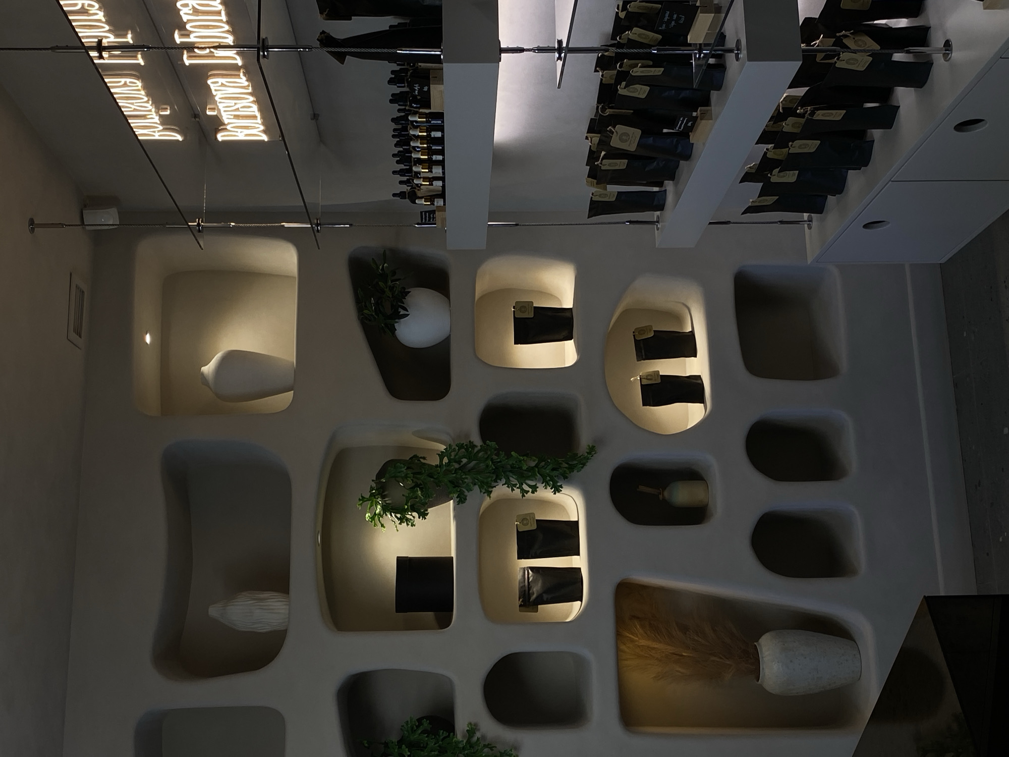 The image depicts an interior wall with irregularly shaped recesses containing various objects. Some recesses hold plants, while others contain what appear to be bags or packages. The wall is painted in a light, neutral color.
Above the wall, the ceiling features a display of wine bottles and packages suspended from a metal frame. A reflective surface above this display shows a neon sign reading "Borisova Loboda."
Based on the sign, this image is from the restaurant "Borisova Loboda" in Izhevsk, Russia.