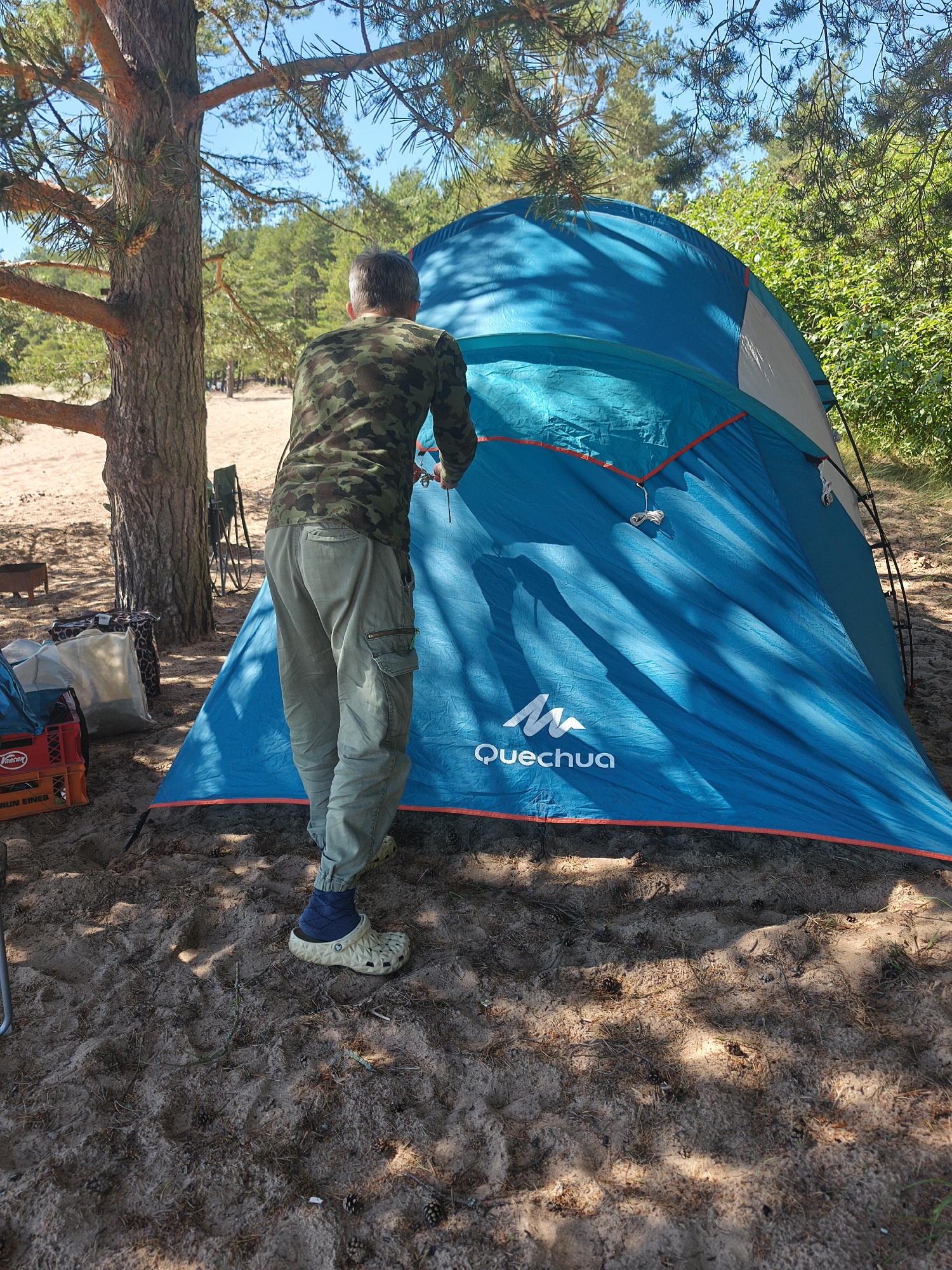 A man, seen from the back, is adjusting a blue tent at a campsite. He is wearing a camouflage long-sleeved shirt, khaki cargo pants, dark blue socks, and beige Crocs. The tent is large and turquoise with an orange trim and a light gray section near the top. The Quechua logo is visible on the tent.
The scene is a sandy area with scattered pine needles and pine cones on the ground, suggesting a forest or wooded location. A large pine tree is to the left of the frame, casting shadows. In the background, there are more trees and green foliage, indicating an outdoor setting, likely in a natural park or a forest campground. To the left of the man, there are several camping items including a red crate with white lettering that reads "Vaalser" and "BRUN EINES", and a couple of patterned bags. A metal grill or fire pit is also visible in the distance.
The time of day appears to be daytime, with bright sunlight creating distinct shadows on the ground and the tent. The weather seems to be clear and sunny. The man's posture suggests he is engaged in setting up or securing the tent. The overall atmosphere is one of leisure and outdoor activity. The location is identified as Zelenaya Roshcha, Russia.