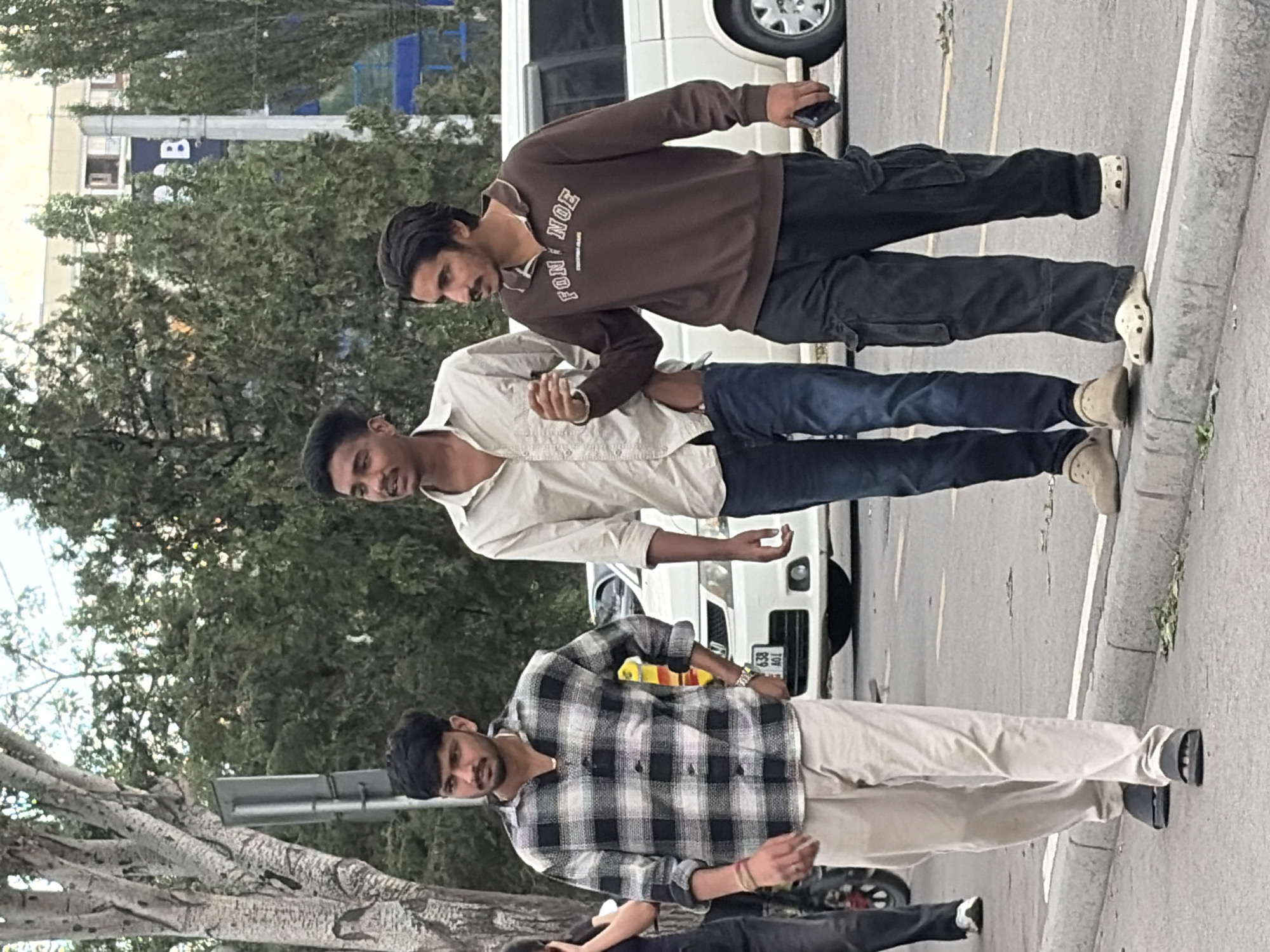 Three young men are walking on a city street. The man on the left wears a black and white plaid shirt and light-colored pants. The middle man is in a light-colored collared shirt and blue jeans. The man on the right is dressed in a brown long-sleeved shirt with lettering and black cargo pants. They are walking on a paved road next to a curb. A white van is visible behind them, as are trees and buildings in the background. The sky is overcast.