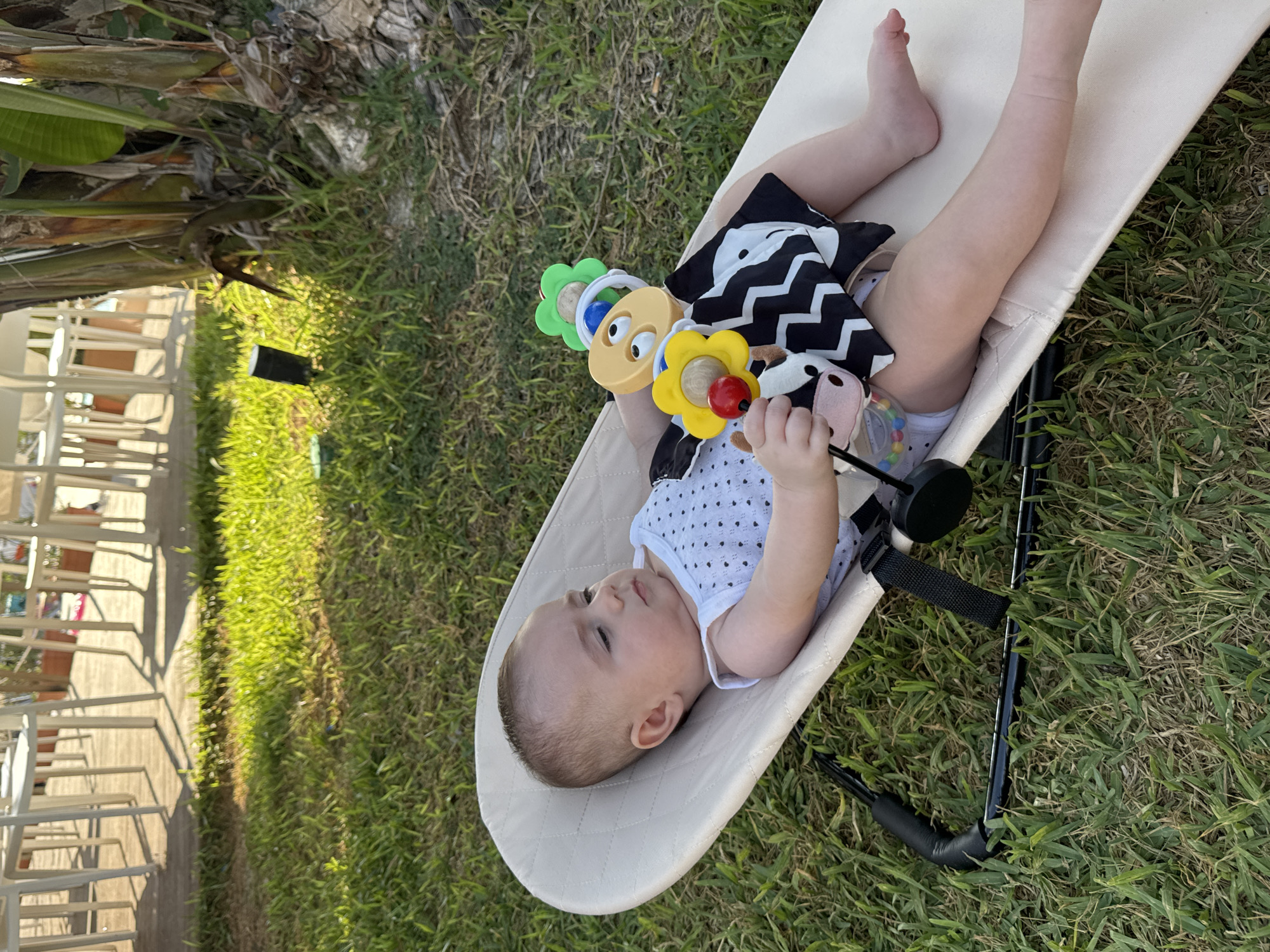 A baby is lying on a beige, fabric infant seat outdoors on a grassy lawn, with a plastic toy in its hands. The baby is wearing a white tank top with black polka dots and a white diaper. The toy is a colorful rattle with different textured parts. In the background, white chairs and tables are visible on a patio area, along with a large plant with broad green leaves. The scene appears to be in Capaci, Italy.