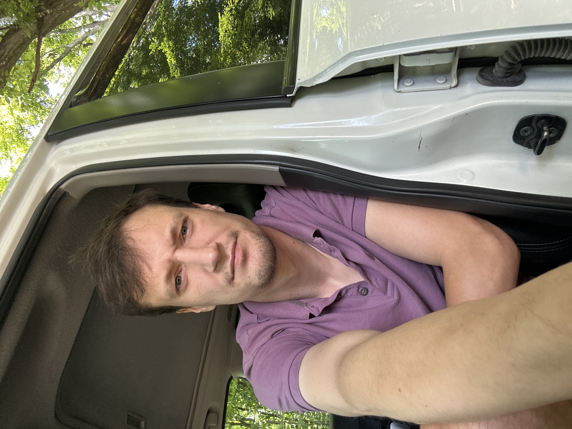 A man with short brown hair and a light complexion is sitting in the driver's seat of a white vehicle. He is wearing a purple polo shirt. His face is turned towards the camera, and he has a neutral expression. The man's left arm is extended forward, resting on his knee or the dashboard. The vehicle's interior, including the roof lining and the door frame, is visible. The open car door reveals a background of lush green trees and foliage, suggesting an outdoor, possibly rural setting. The specific location of Shatava, Ukraine, is not discernible from the image alone.