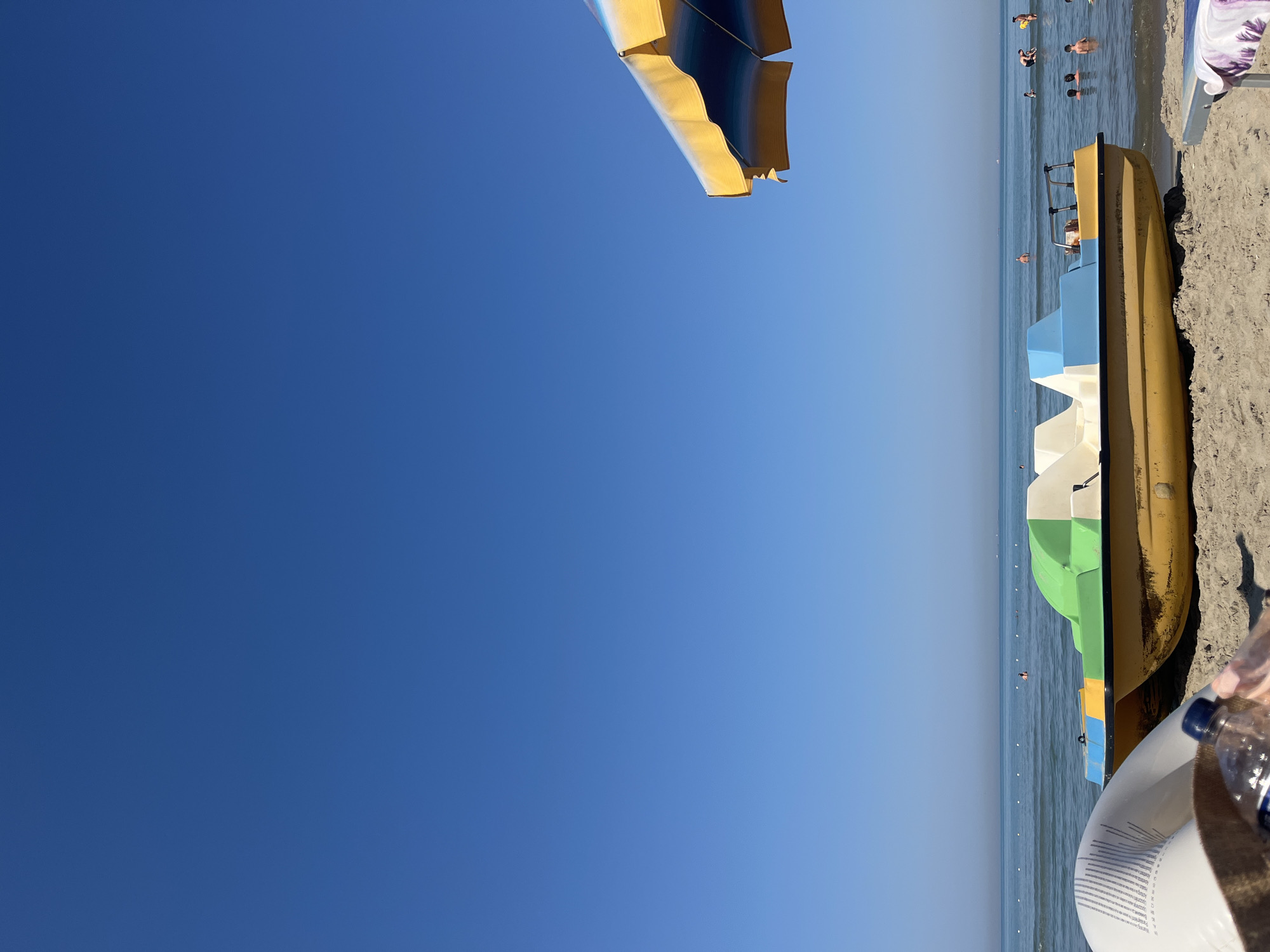 The image depicts a sunny day at a beach in Bashkia Durrës, Albania. In the foreground, a yellow and white pedal boat with green and blue accents rests on the sand near the water's edge. An inflatable ring and a water bottle are also visible on the sand in the lower left portion of the frame. Beyond the pedal boat, the calm sea stretches towards the horizon, with a few people swimming and wading in the shallow water. A beach umbrella with yellow and blue stripes is partially visible in the upper right corner. The sky is a clear, vibrant blue, indicating good weather conditions.