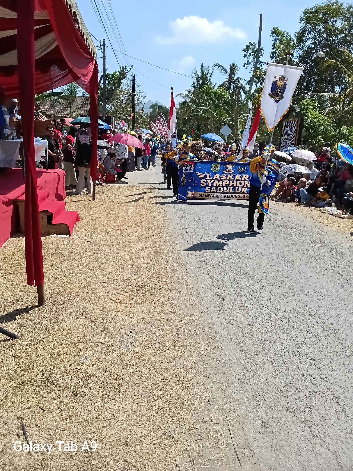 A daytime outdoor scene depicts a parade on a paved road. In the foreground, a red and white tent structure is partially visible on the left, with some dry straw-like material covering the ground. Further down the road, a group of people in blue and gold marching band uniforms are parading, carrying a banner that reads "LASKAR SYMPHON SADULUR SD NEGERI 1 KEDUNGGLUR". One individual in the band is holding a white flag with a crest and gold trim. Spectators, some holding umbrellas, are seated and standing on the right side of the road, observing the parade. The background features trees and houses under a clear blue sky with some clouds. The image is labeled "Galaxy Tab A9" in the lower left corner. This event appears to be taking place in the village of Ngadirenggo, Indonesia, given the text on the banner.