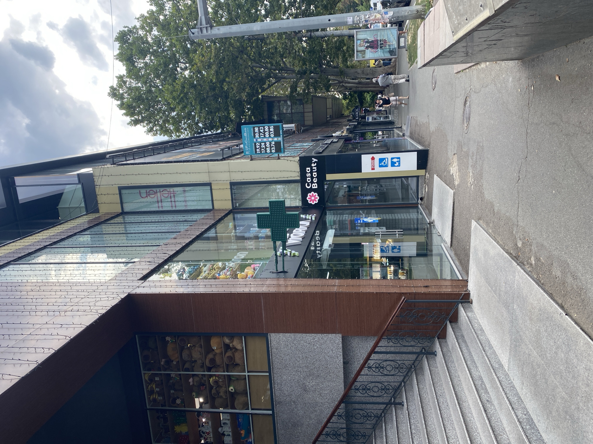 The image captures a street scene in Chișinău, Moldova, specifically on a commercial building's ground floor. The building features a modern facade with large glass windows, revealing shops and displays inside. One storefront clearly visible is "Casa Beauty," adorned with a pink floral logo. Adjacent to it, a green cross symbol indicates a pharmacy. A digital display board showing currency exchange rates for FinComBank is mounted on the building's exterior. In front of the shops, a paved sidewalk runs alongside the street, with a few people walking. A set of granite stairs with a decorative metal railing leads up to an entrance on the left side of the frame, with a ramp running alongside it for accessibility. The street is lined with trees, and the sky is partly cloudy. The overall impression is a typical urban commercial area.