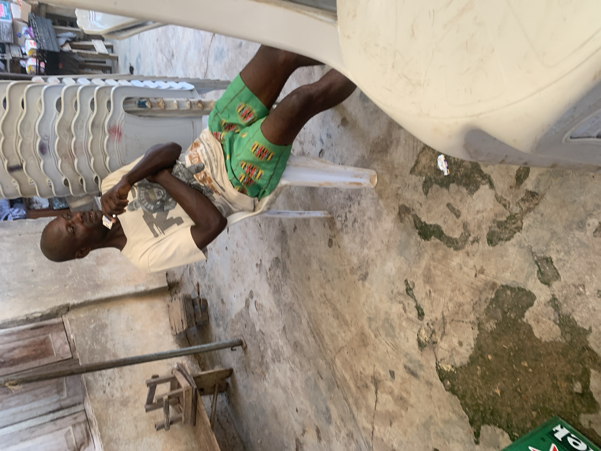 A black man sits on a white plastic chair in what appears to be an outdoor, possibly informal, gathering space in Shagamu, Nigeria. He is wearing a light-colored t-shirt with a graphic print and green shorts with a colorful pattern. He is holding a small white object, possibly a piece of candy or a wrapper, in his right hand and appears to be looking at it. His arms are crossed, and he is leaning back slightly in the chair.
The environment is somewhat rustic. To the left of the man, there is a wooden bench with a small wooden stool on top of it. Behind the man, a stack of white plastic chairs is piled high. The floor is concrete and shows signs of wear, with patches of dampness and dirt. A portion of a green Heineken bottle is visible in the lower left corner of the frame, suggesting the presence of beverages. The background wall is textured and appears to be made of concrete or plaster, with some wooden elements visible. The overall impression is a casual, everyday scene in a Nigerian setting.