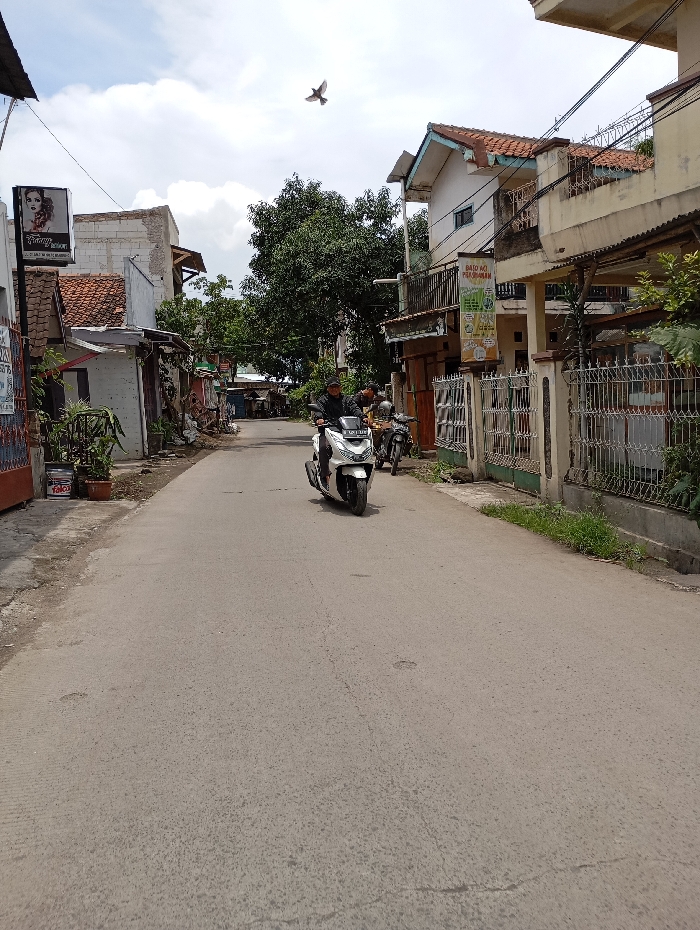 The scene is a street in Katapang, Indonesia, located at -6.985 latitude and 107.557 longitude. It's a residential street with buildings on both sides. There is a man riding a white scooter in the middle of the street. The scooter is moving towards the right side of the image. There is a second scooter parked on the right side of the image with a man sitting on it. The man on the scooter is wearing a helmet and a white shirt. The road is paved and empty, except for the scooter. The sky is clear with some clouds. A bird is flying in the sky. The surrounding buildings are residential, and some have metal fences. There is a sign on the left side of the image that says "Hair Salon". There is a sign on the right side of the image that says "DATO AC PALOMA". There is a tree on the left side of the image, and another tree on the right side. There are some plants growing on the side of the road. There is a white building on the right side of the image with a large metal fence. There is a small group of buildings in the left side of the image, appearing to be a commercial area. The street appears to be relatively quiet.