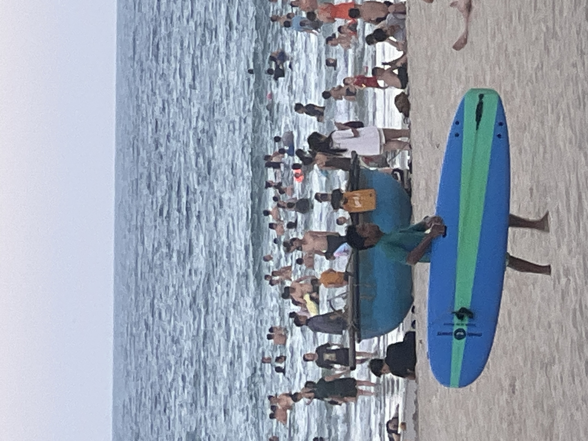 The image depicts a crowded beach scene at the city of Đà Nẵng, Vietnam. In the foreground, a young boy in a green t-shirt carries a blue and green surfboard. Behind him, several people are visible on the sandy beach, including a person lying down. The water is filled with numerous swimmers of all ages, enjoying the ocean. Several boats are present near the shore. The sky is overcast, and the ocean is calm with gentle waves. The overall atmosphere is lively and suggests a popular recreational area.