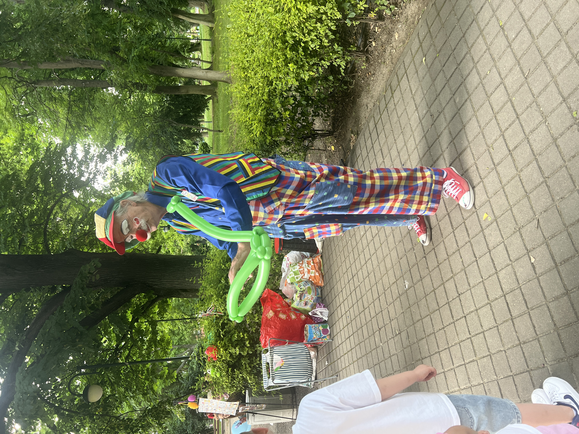 A clown is making balloon animals in a park in Plovdiv, Bulgaria. The clown, wearing a blue shirt, colorful plaid pants, a multi-colored striped vest, a red nose, and red sneakers, is bending over a green balloon. Next to him is a folding chair with various bags and items on it. The ground is paved with gray square tiles. Trees and bushes form a green backdrop. Part of a person wearing a white shirt and blue shorts is visible in the lower right corner.
