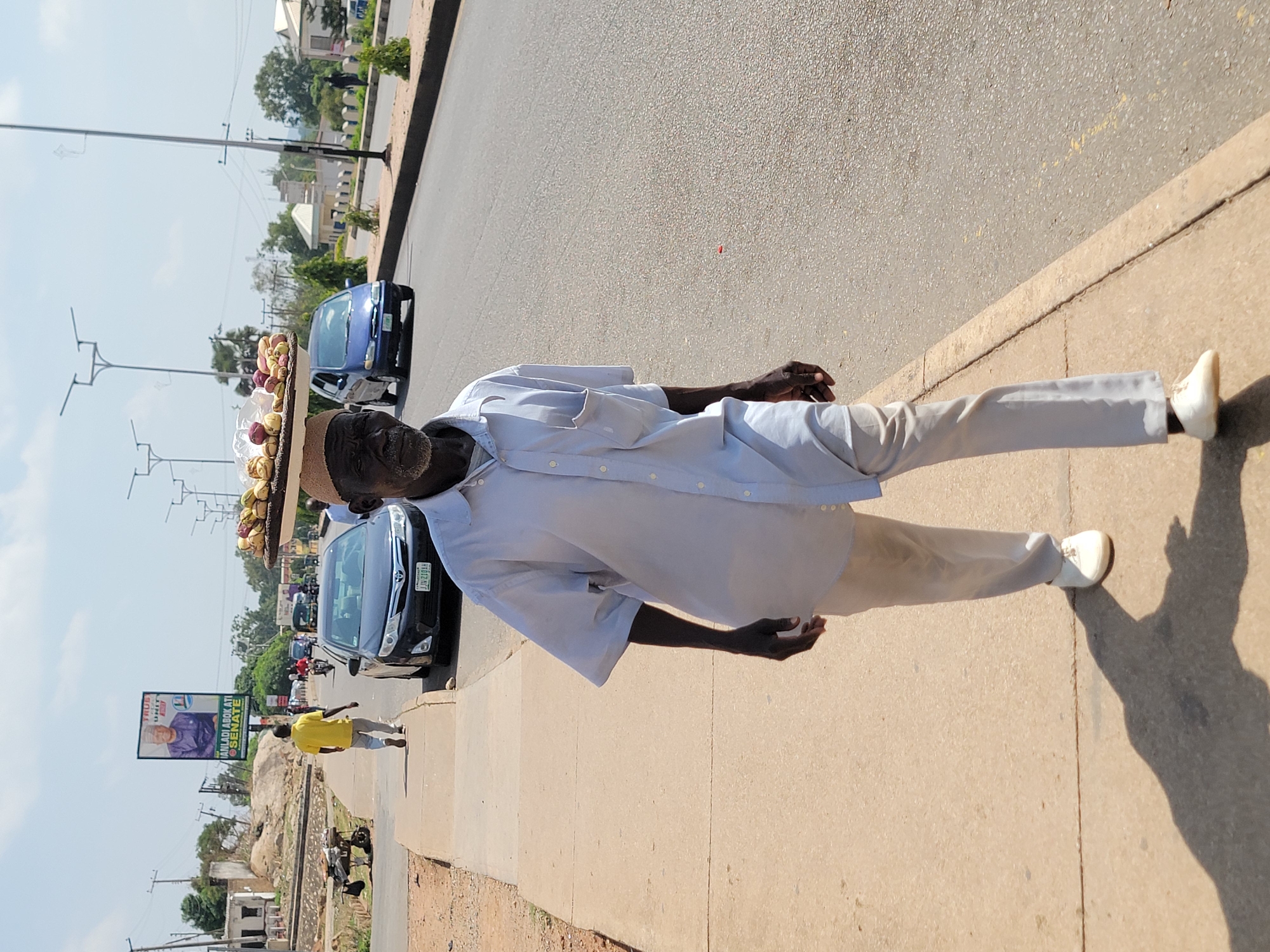 An elderly African man walks along a sidewalk in Jos, Nigeria. He carries a tray of goods on his head, likely for sale. He is wearing a light-colored button-down shirt, light-colored pants, and white shoes. The sidewalk runs adjacent to a busy street with moving cars. A billboard featuring a politician is visible in the background. In the distance, a building can be seen along the horizon.