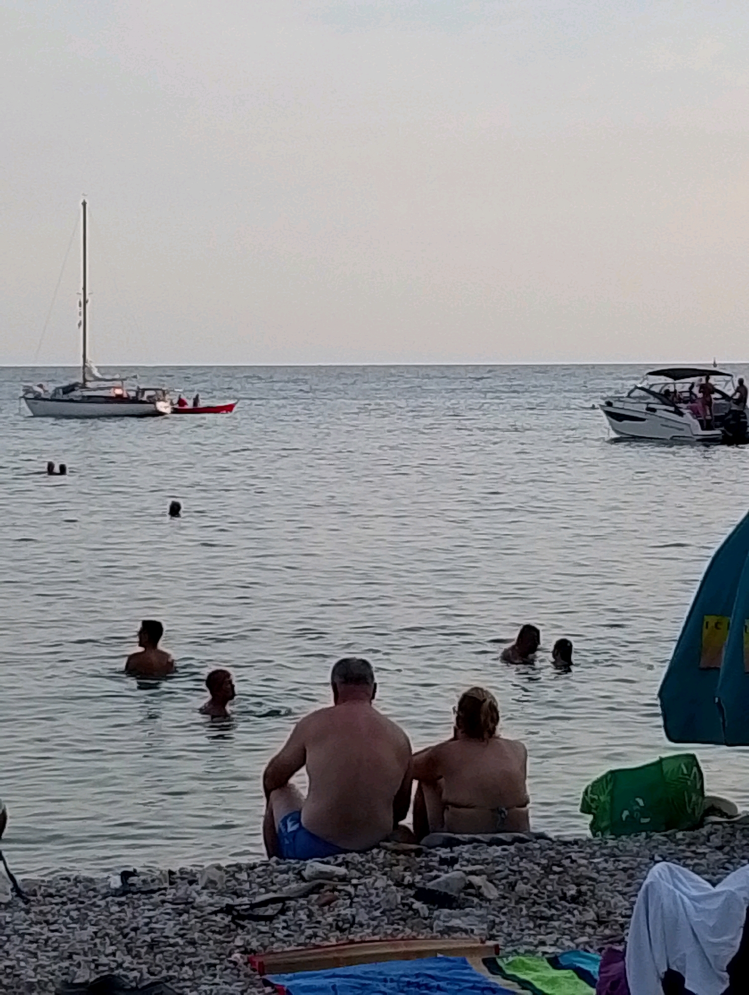 A wide shot captures a tranquil scene at the Sutomore, Montenegro coastline during dusk. The sky is a muted, pale gray, hinting at the approaching evening. The sea is calm with gentle ripples, reflecting the soft, diffused light.
In the foreground, a couple sits on a pebble beach, facing the water. The man, on the left, wears blue swimming shorts and is seen from behind. The woman, to his right, also facing away from the camera, is in a bikini.
The water is populated by several individuals. To the left of the couple, a group of people are partially submerged, swimming or wading. Further out, a sailboat with a white hull and a red inflatable dinghy attached is anchored, with several figures visible on deck. To the right of the sailboat, a modern motorboat is underway, carrying multiple passengers.
Scattered across the water are other swimmers, their heads and shoulders visible above the surface. On the beach, a green bag and various beach towels are visible, along with the corner of a blue beach umbrella, suggesting a relaxed beach setting. The overall atmosphere is one of quiet leisure as the day winds down.