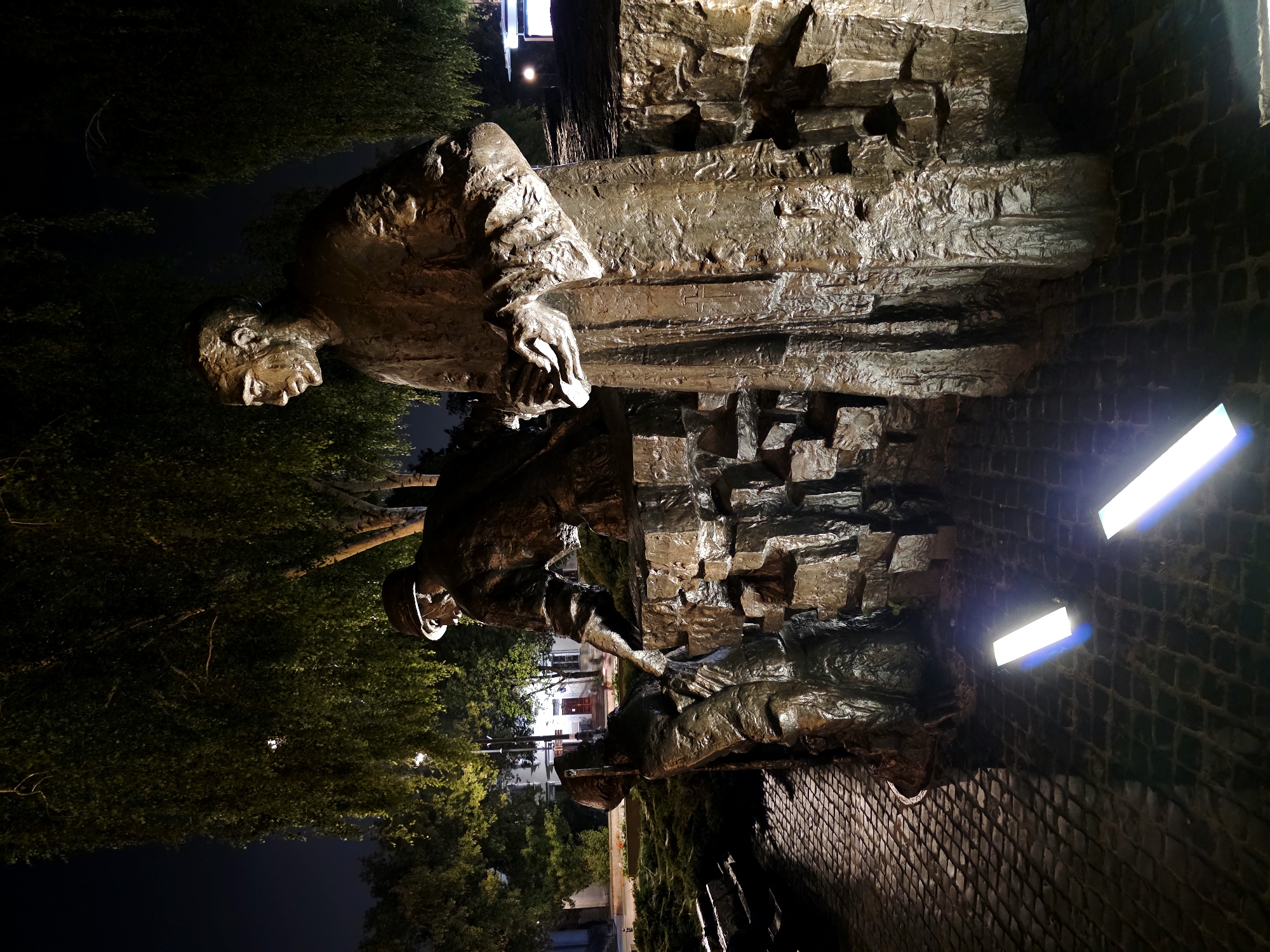 This is a nighttime, eye-level photograph of a bronze sculpture depicting men assisting each other. The sculpture is located in Warsaw, Poland, near the church of St. Stanislaus Kostka in the Żoliborz district, commemorating victims of the communist regime.
In the foreground, a large, standing figure in a robe, possibly a clergyman, extends his hands forward. To his left, a second figure, wearing a helmet and a soldier-like uniform, is crouched and reaching out to assist a third, smaller figure who is kneeling or falling. The figures are interacting with each other in a scene of mutual aid or rescue.
The sculpture is placed on a textured, rocky base, and behind it are trees with lush foliage. The ground in front of the sculpture is paved with cobblestones, and two rectangular lights are embedded in the ground, casting bright beams upwards. The background is dark, with some ambient streetlights and the outline of buildings visible in the distance. The overall mood is somber and reflective, enhanced by the dramatic lighting of the night scene.