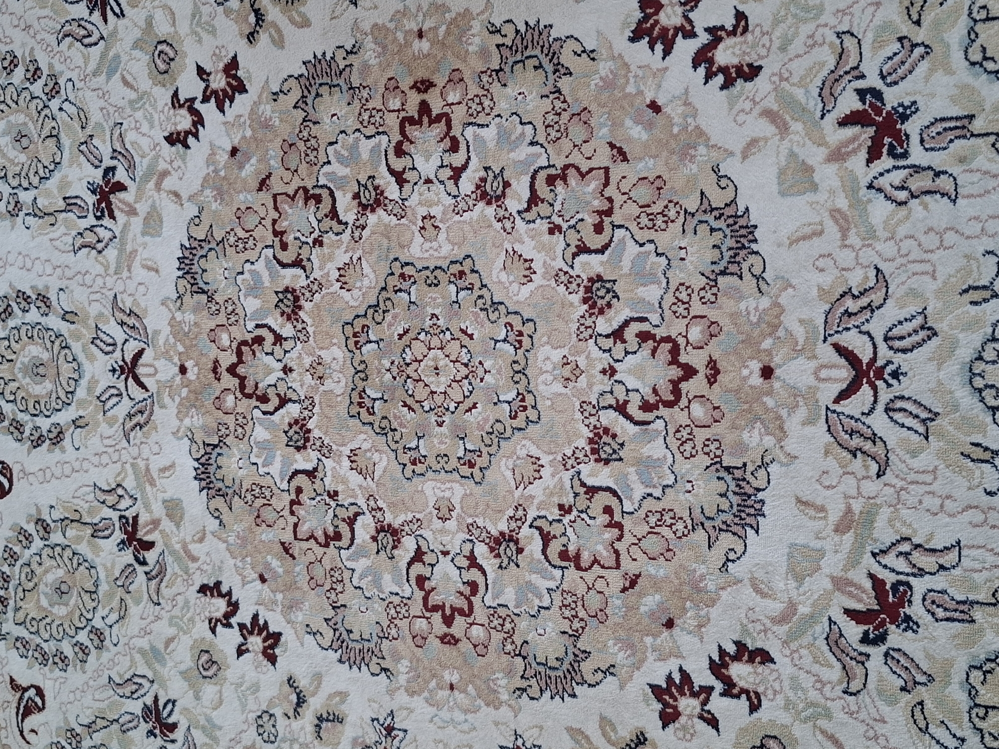 The image shows a close-up of a white rug with a floral pattern. The pattern features intricate designs in shades of red, beige, and muted blue. The rug is likely indoors, as indicated by its texture and the absence of outdoor elements. There are no people or any discernible location in the city of Almaty, Kazakhstan visible in the image. The focus is entirely on the textile design of the rug.