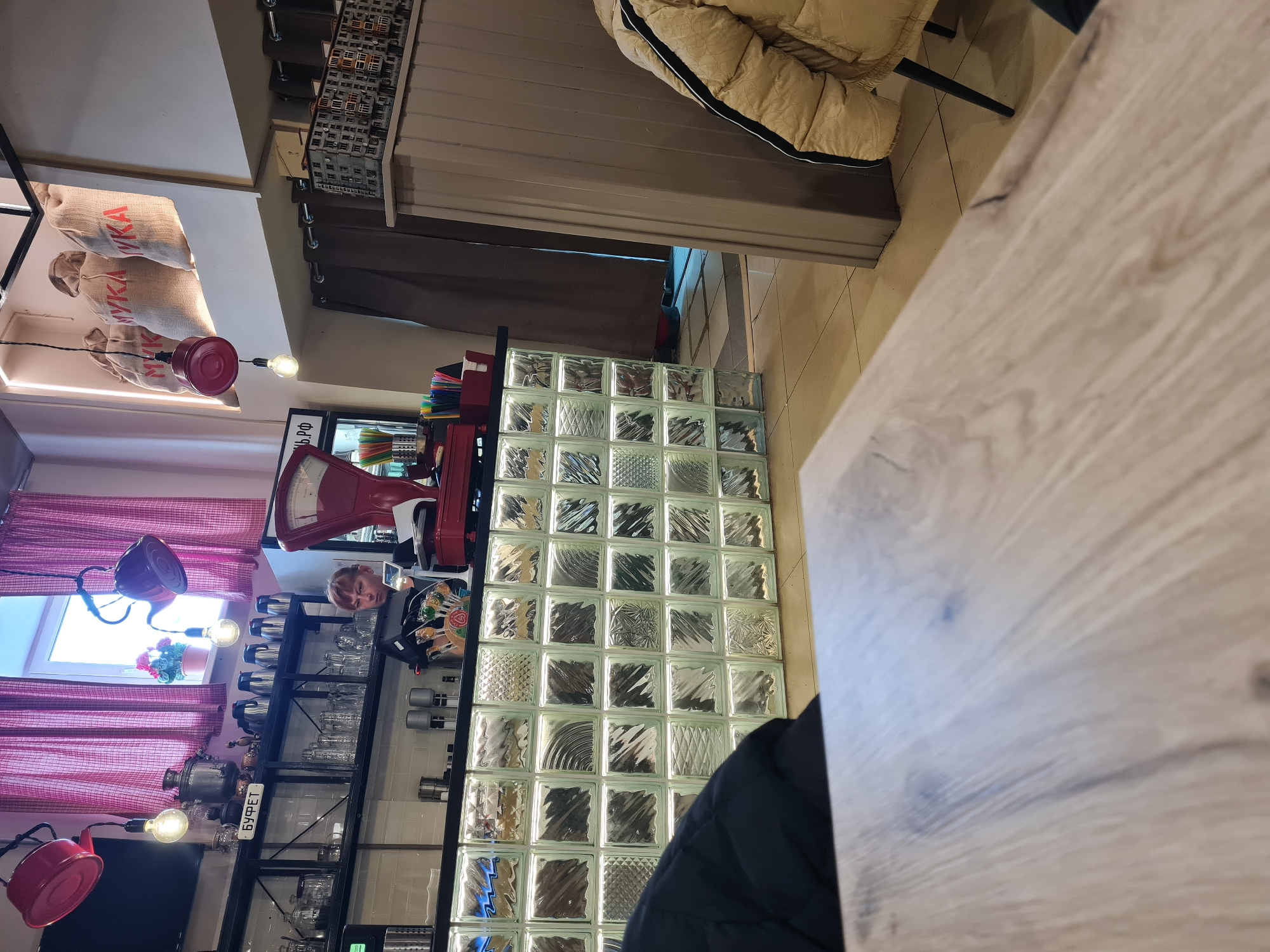 The scene depicts a section of a cafe or restaurant interior in Izhevsk, Russia. A blonde-haired child stands behind a glass block partition, next to a red vintage scale and a shelf displaying glassware and samovars. Red lamps hang above the shelf, and red-and-white striped curtains adorn a window. In the background, bags labeled "MYKA" are visible on a shelf. In the foreground is a wooden table surface and the edge of a dark jacket. The background shows a hanging light with multiple bulbs. The overall impression is that of a quaint, possibly themed establishment, with the glass brick wall acting as a partial divider.