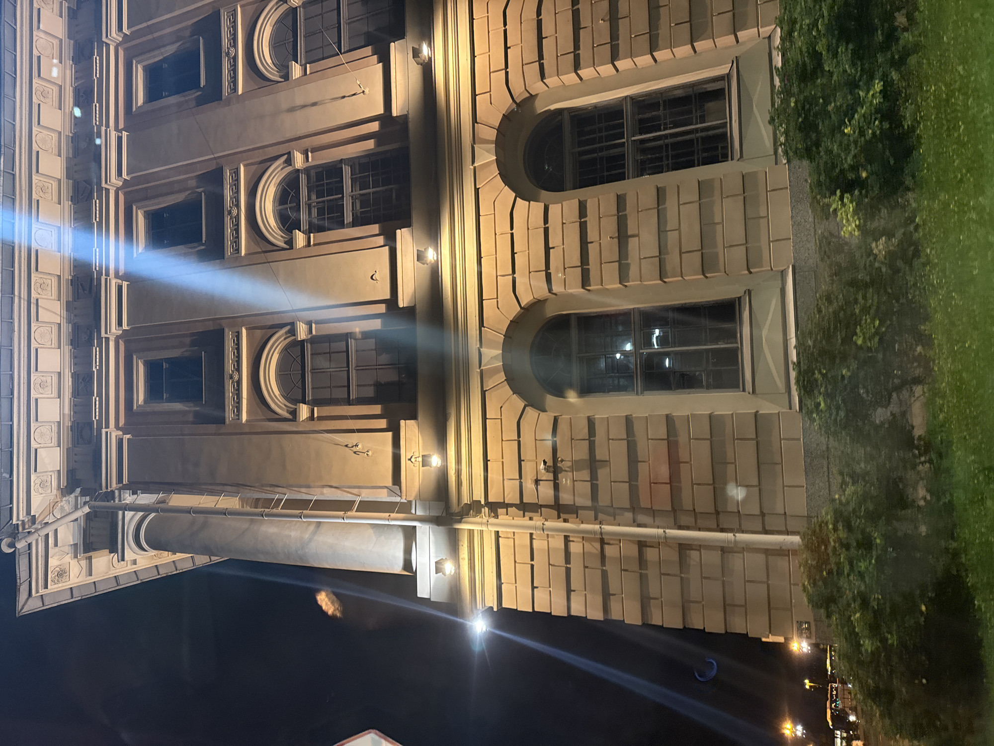 The image captures a nighttime view of a building with classical architectural elements in Saint Petersburg, Russia. The building is characterized by its light-colored stone facade, tall windows with arched tops on the lower floors, and smaller rectangular windows on the upper floors. Decorative friezes and cornices are visible along the roofline and between the window levels. A large, fluted column is present on the left side of the building. A white pipe runs vertically down the facade. Lighting from street lamps and possibly from within the building casts a warm glow on the structure. In the foreground, there are green bushes and grass, suggesting a street or park setting. The sky is dark, indicating it is nighttime. There are no visible people in the image. The overall impression is of an imposing and historic structure illuminated at night.