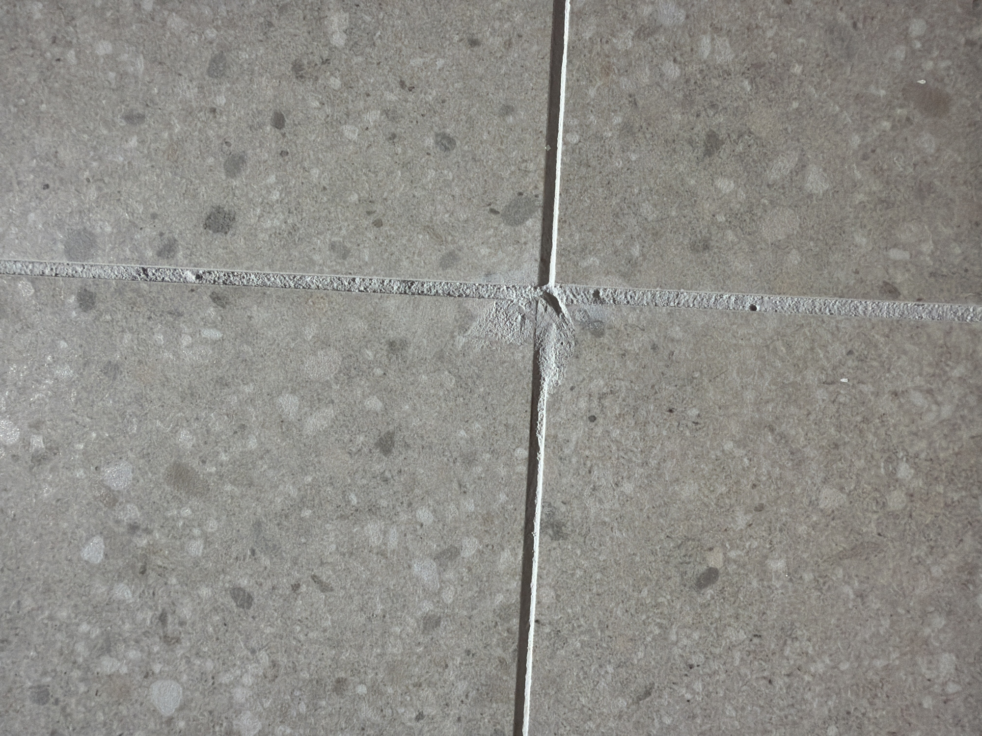 The image displays a close-up, overhead view of two grey ceramic tiles meeting at a grout line, with another horizontal grout line intersecting them. The tiles have a speckled pattern with variations in tone, appearing to be made of a textured material. The grout is lighter in color and shows some signs of wear or chipping, particularly at the central intersection. The lighting suggests an indoor or shaded outdoor environment, casting subtle shadows. There are no people or other discernible objects in the image, focusing solely on the texture and arrangement of the tiles. This image does not contain any information that would allow for its location to be identified as Jerusalem, Israel.