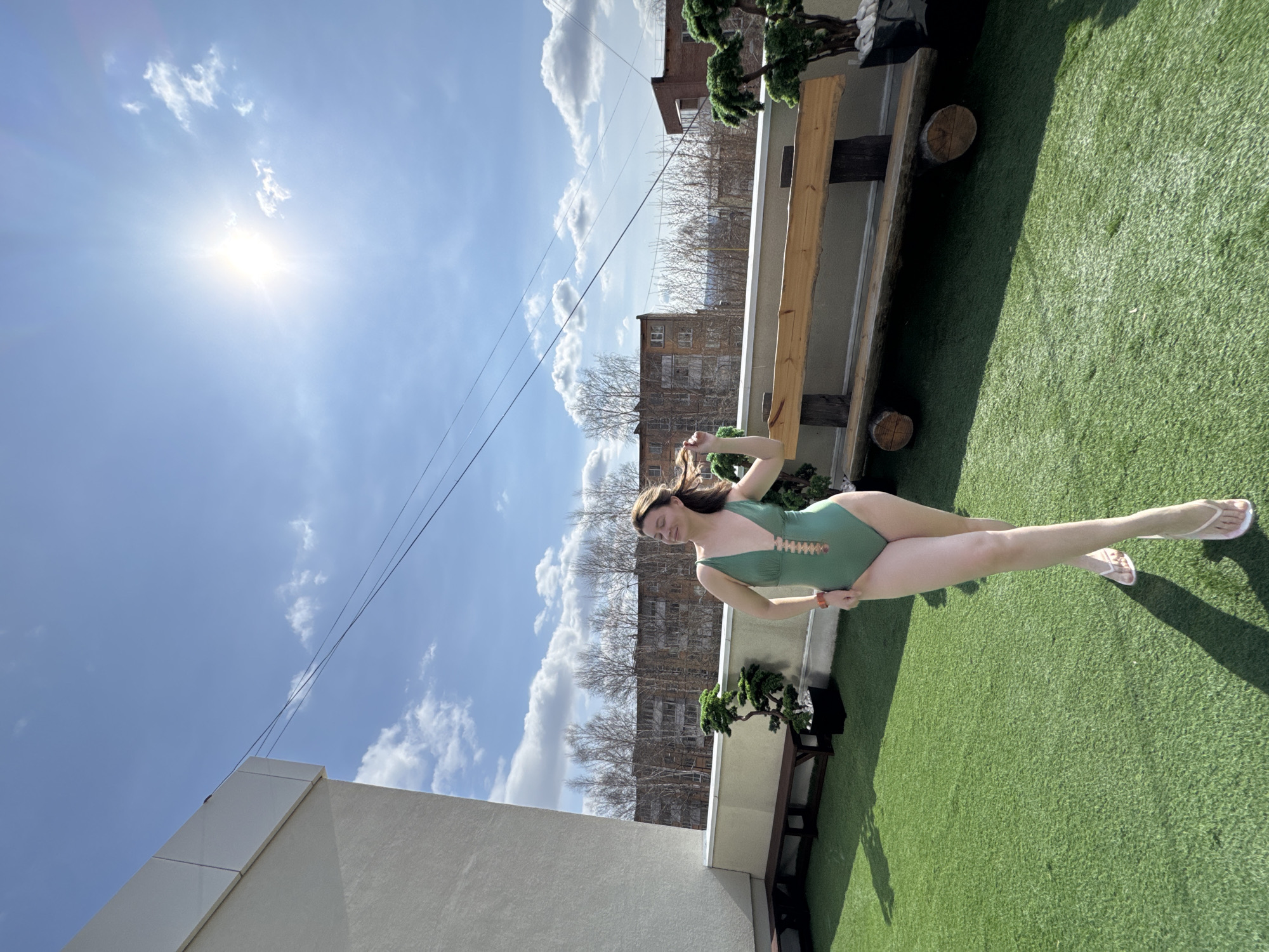 A woman is reclining on a rooftop covered with artificial turf. She is wearing a green swimsuit and white sandals. Her left arm is raised, and she is touching her hair. A wooden bench and a small potted tree are behind her. In the background is a building with a row of bare trees in front of it. The sky is blue with scattered clouds, and the sun is visible in the upper left corner of the frame. Power lines are also visible. The image was probably taken in Votkinsk, Russia.