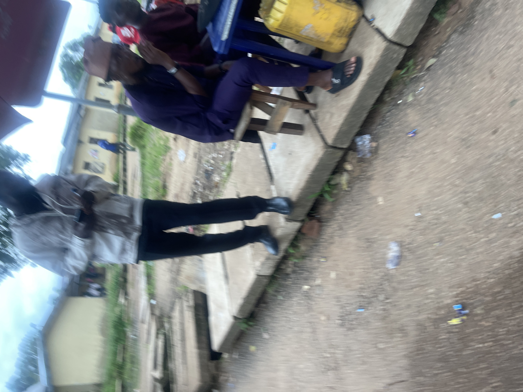The image depicts an outdoor scene with several individuals. A person in a white jacket and black pants stands on a concrete structure, holding a phone. To their right, another person wearing a purple outfit and a cap sits on a small wooden stool. Further to the right, a person in a maroon outfit is visible, partially obscured by a blue table. A large yellow container is situated next to the table. The ground appears to be a wet, concrete surface with some debris scattered around, including plastic bottles and wrappers. In the background, there are buildings and some greenery, suggesting an urban or semi-urban environment. The overall atmosphere is casual, possibly a street-side gathering or a market area. The exact location of Rafin Par, Nigeria cannot be confirmed from the visual information alone.