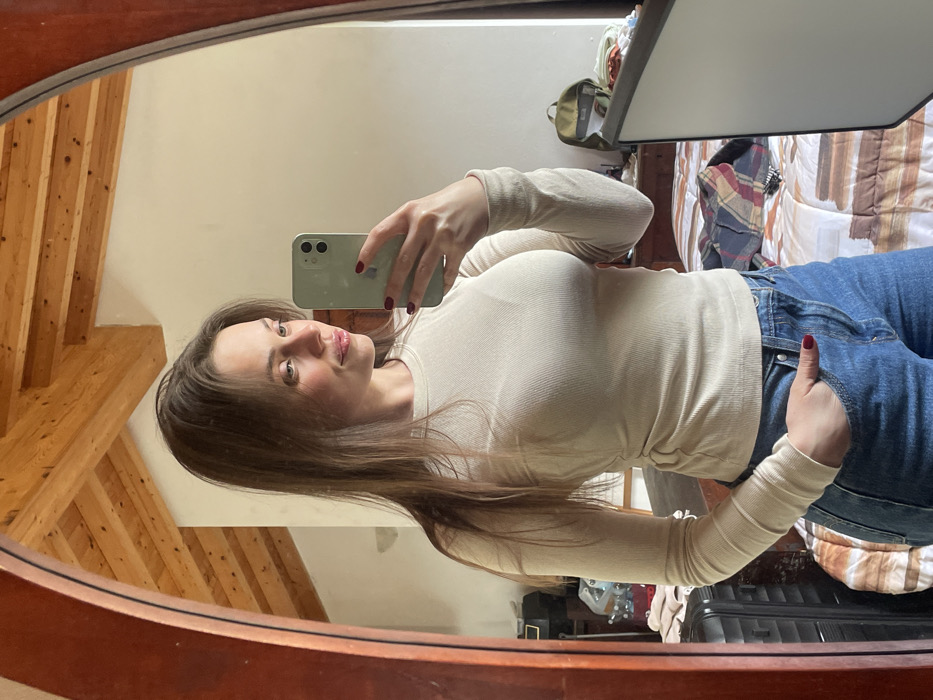 A female subject is depicted taking a mirror selfie indoors in a residential space located in Monsummano Terme, Italy. The subject, a young female with long brown hair, is shown from the waist up, wearing a light beige, long-sleeved ribbed top and blue denim jeans. She holds a light green smartphone with her left hand, featuring red-painted fingernails, positioned to capture her reflection. Her right hand is partially visible, resting in the pocket of her jeans. The subject displays a subtle smile and gazes towards her left. The environment reflected in the large, wooden-framed mirror reveals a room with an exposed wooden beam ceiling and light-colored walls. Visible background objects include a bed with a brown and white striped comforter, a plaid shirt draped on the bed, a green backpack, and a black hard-shell suitcase.