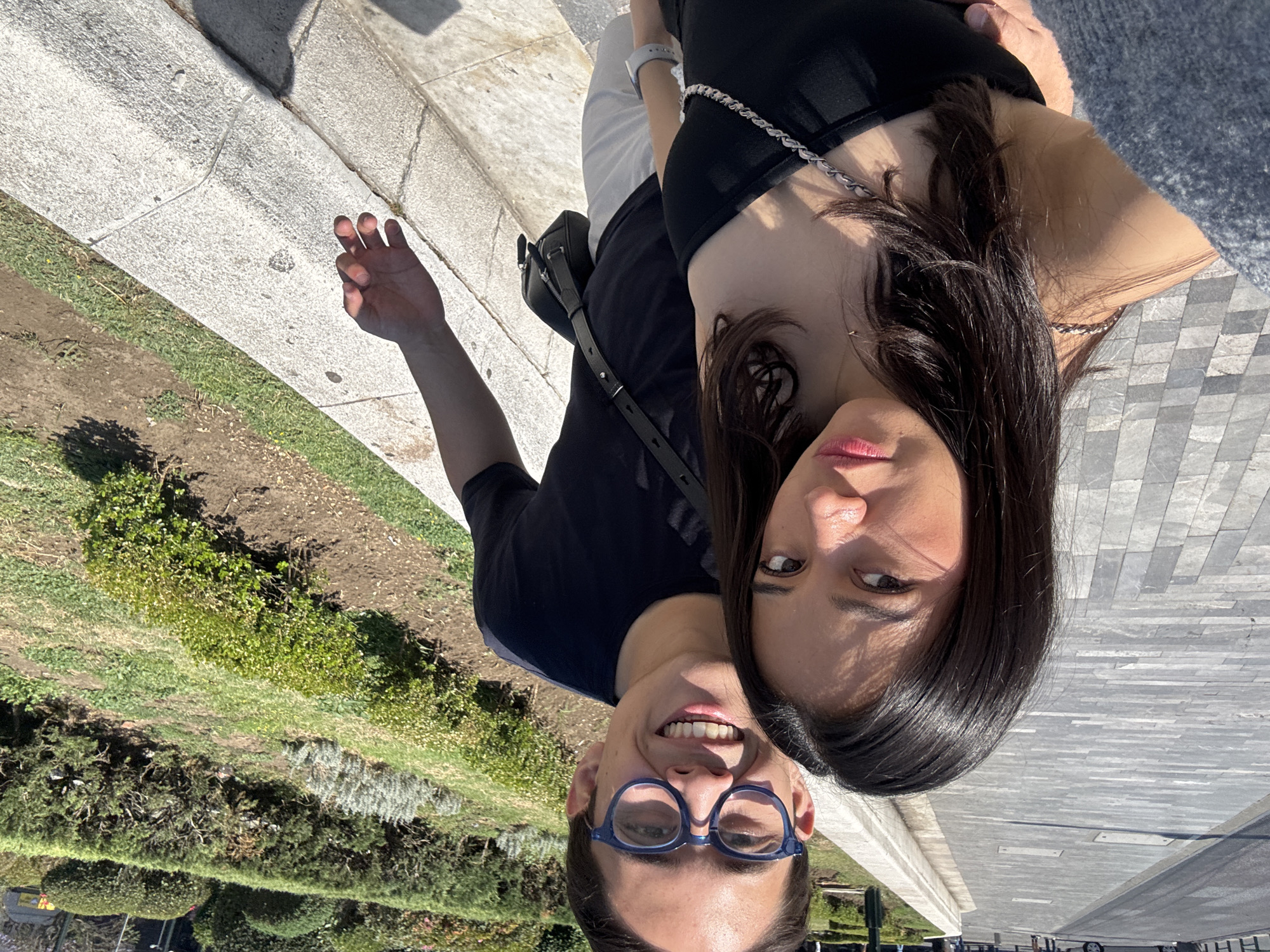 A man and a woman are captured in a close-up selfie. The man is wearing glasses and smiling. The woman is beside him. The background features greenery, a paved area, and what appears to be part of an urban landscape.
The location appears to be Mexico City, Mexico.