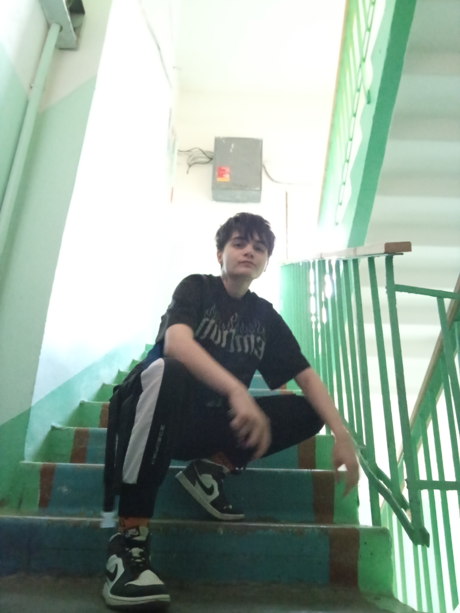 A young person with short, dark hair is crouching on painted concrete stairs with green railings. The person is wearing a black t-shirt, black pants with a white stripe, and black and white high-top sneakers. The stairs are painted teal with brown edges. A grey electrical box is visible on a white wall in the background. The scene appears to be a stairwell of a building. The location is identified as Naberezhnye Chelny, Russia.