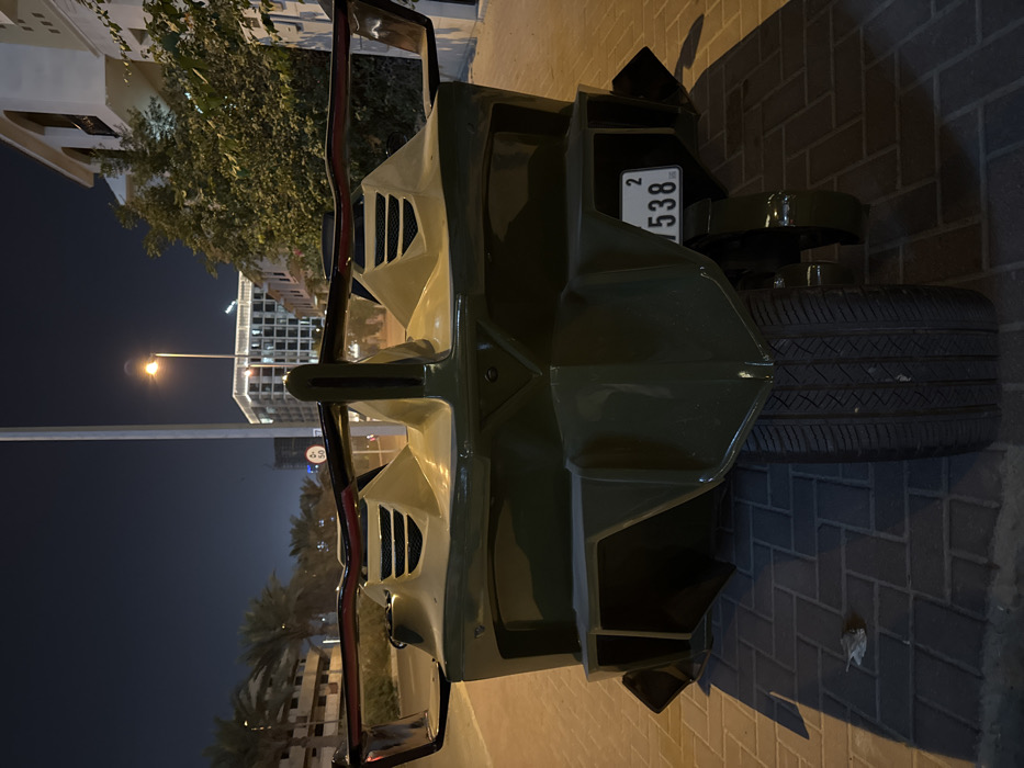 Centered is a military-green vehicle with a spare tire mounted on the rear. A license plate displaying "5382" is visible. The vehicle is parked on a brick-paved surface, likely a sidewalk or street. The background features buildings, including one that appears to be residential, with palm trees in the distance. A street light illuminates the nighttime scene.