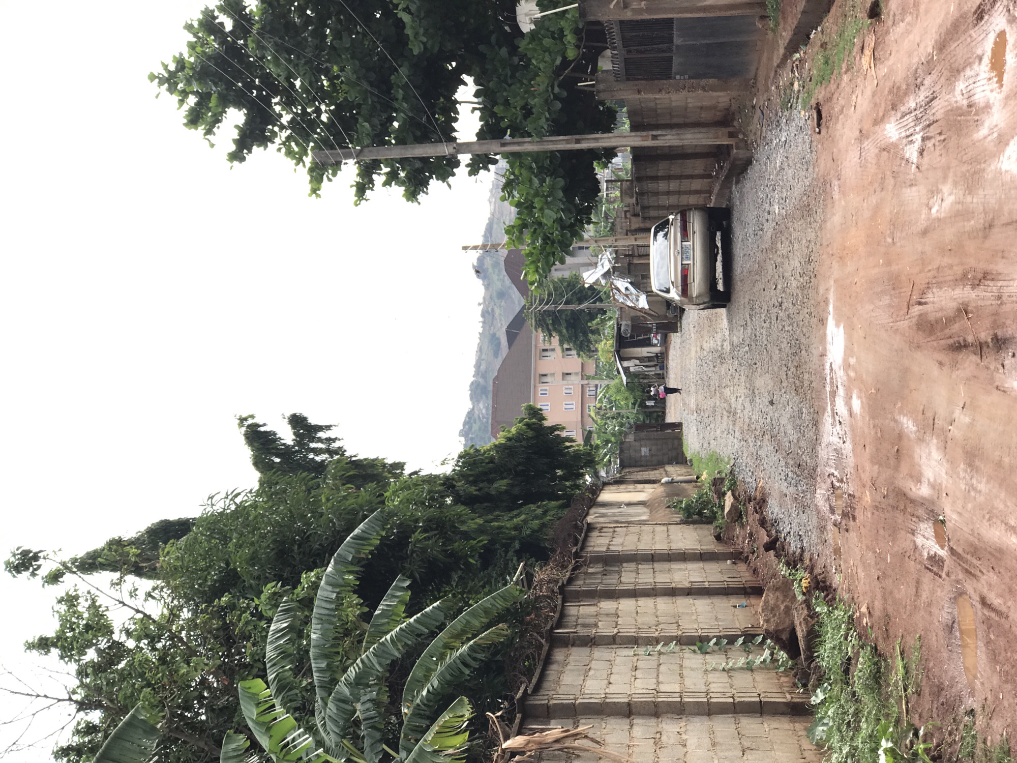 The image depicts a street scene in Dutsen Alhaji, Nigeria. A car is parked on a gravel section of the road. The road surface transitions to mud with visible tire tracks and puddles. A tall cement wall and vegetation line the left side of the frame. In the background, buildings are visible on a hill. An electric pole is seen on the left. Two figures are seen walking in the distance. Banana trees are visible on the left.