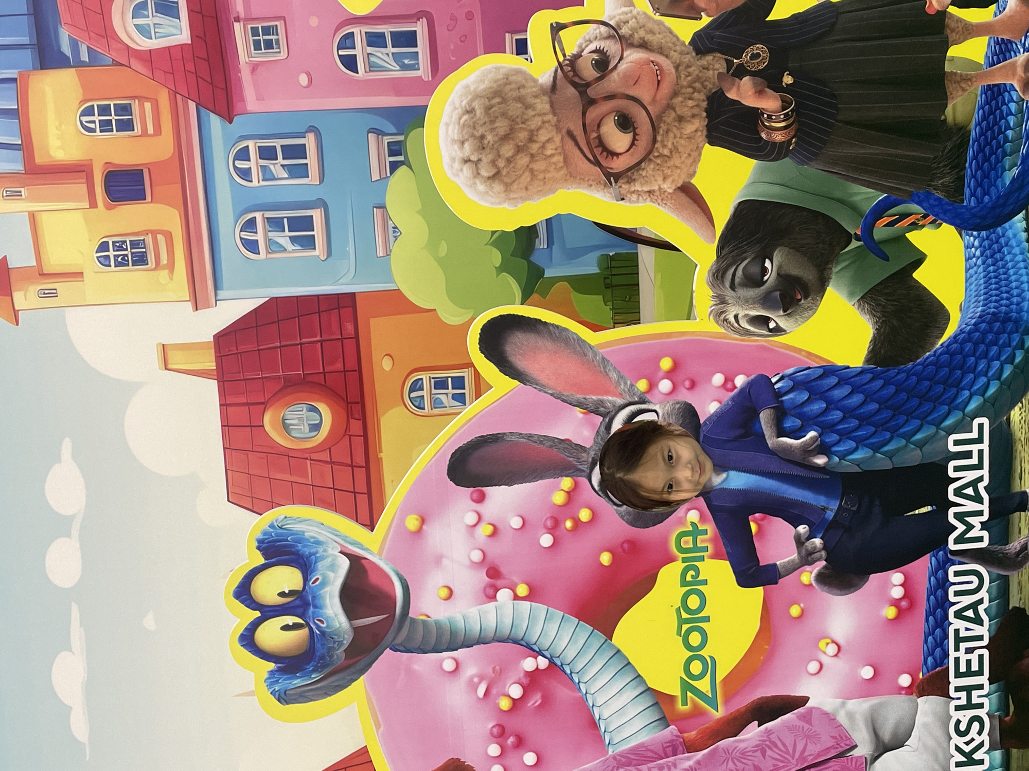This image is a photograph of a large, colorful advertisement or backdrop featuring characters from the Disney movie "Zootopia." The advertisement is located in Kokshetau Mall in Kokshetau, Kazakhstan.
The advertisement depicts several characters: a large blue snake with a gaping mouth, a pink donut with sprinkles, a grey rabbit character with long ears, a sloth character, and a sheep character wearing glasses and a suit. A young girl's face has been superimposed onto the body of the rabbit character. The background of the advertisement is a stylized cityscape with brightly colored buildings. The bottom of the advertisement displays the text "KSHETAU MALL" and "ZOOTOPIA" in large, prominent lettering. The overall impression is of a promotional display for the movie, likely placed in a public area like a shopping mall.