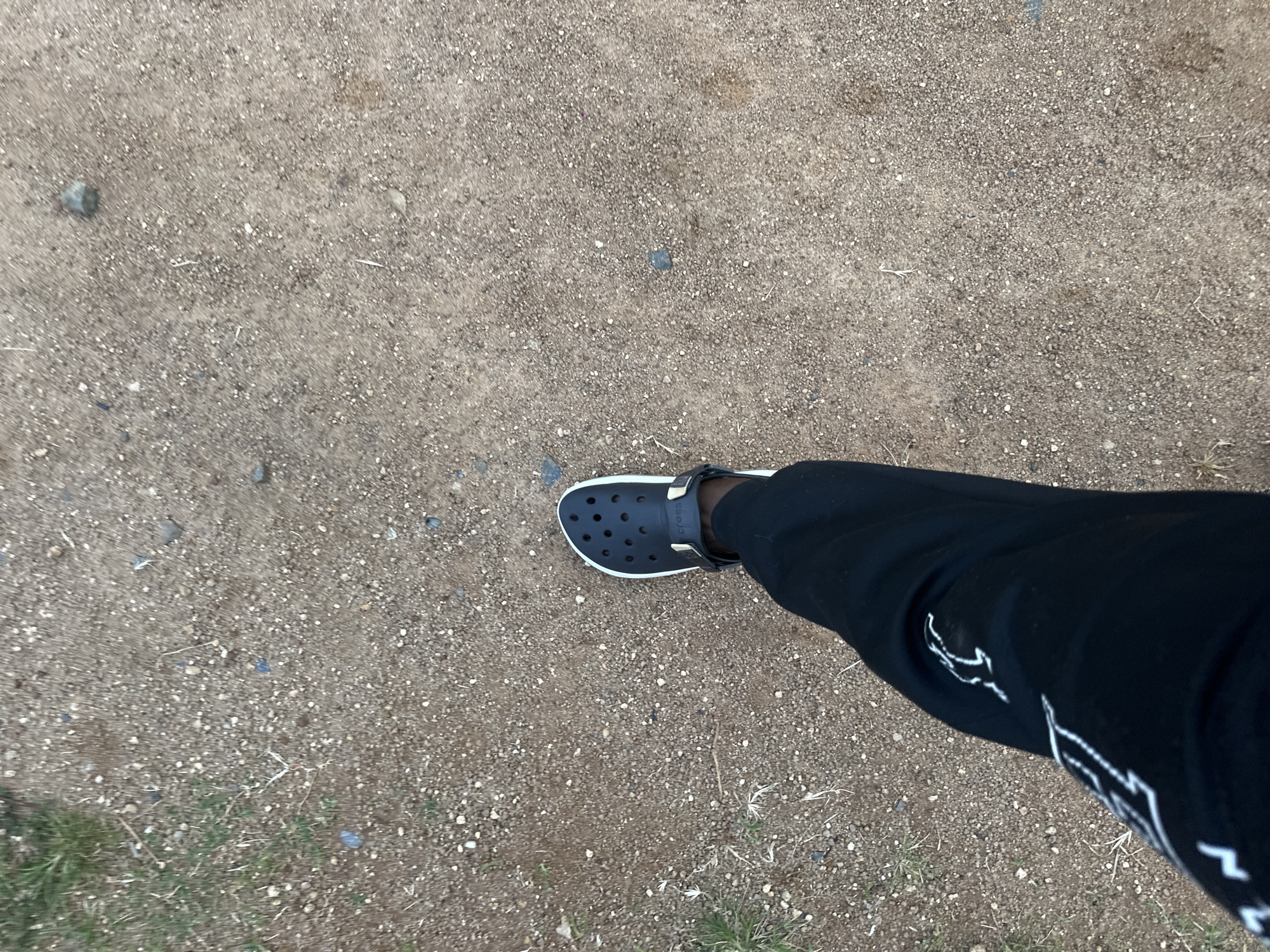 The media file shows a person's leg and foot wearing black Crocs and black pants. The Crocs are dark and appear to have a white trim. The pants are also black and have white graphic designs on them, possibly a logo. The person is standing on a ground covered with small stones and dirt. Some sparse dry grass is visible in the lower part of the frame. The scene appears to be outdoors, given the ground texture and the natural lighting. The time of day is likely daytime, as indicated by the brightness of the scene. The weather seems to be dry, with no signs of rain or wetness. The image provides no clear indication of the specific location within Gwong, Nigeria, beyond it being an outdoor area with a rough, unpaved surface. The activity depicted is simply the person standing or walking, as suggested by the angled perspective of the leg and foot. There is no explicit emotional expression conveyed due to the focus on the lower body and the ground. Some text is visible on the Crocs, appearing as a white logo on the side strap, and also on the pants as white graphic patterns.