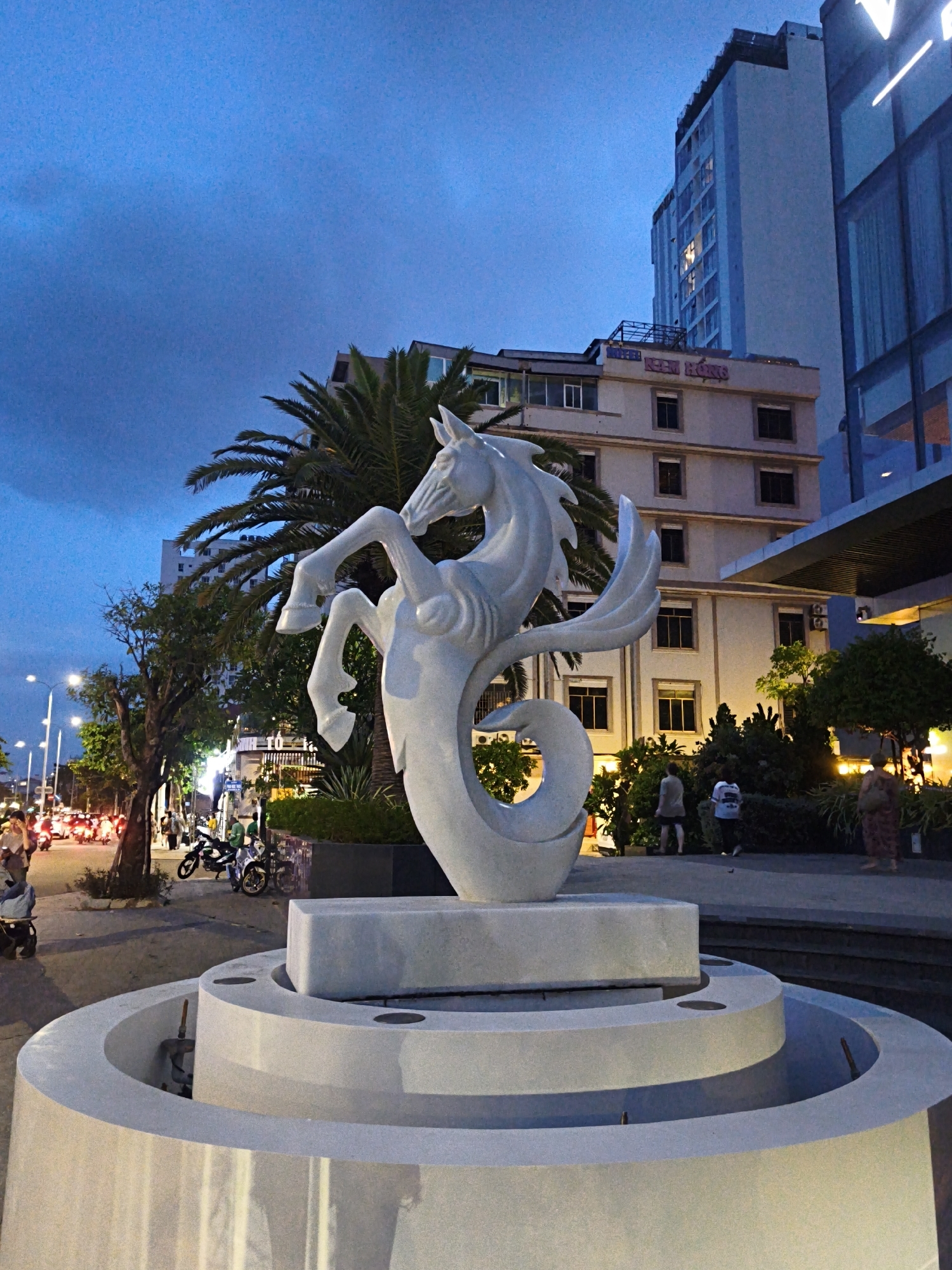 This media file depicts a sculpture of a seahorse or a mythical creature with horse-like features, possibly a hippocampus, in a public outdoor space. The sculpture is white and appears to be made of stone or concrete, mounted on a multi-tiered circular fountain base. The creature is depicted in a dynamic pose, with its front legs raised and its tail curled.
The scene is set at dusk or early evening, as indicated by the dim light and the illuminated streetlights and building windows. The sky is a deep blue, suggesting twilight. In the background, several buildings are visible, including a tall, modern high-rise with lit windows, and a lower, older building with signage that reads "HOTEL KIM HONG".
The surrounding area shows a street with traffic, including many motorcycles and scooters, typical of a Vietnamese urban setting. People are walking on the sidewalks and near the buildings. There is lush greenery, including palm trees and other bushes, around the sculpture and buildings, suggesting a tropical or subtropical location. The overall mood appears to be calm and tranquil, despite the presence of traffic. The location cues, such as the architecture and the prevalence of motorcycles, strongly suggest an urban environment in Vietnam, specifically Bac Nha Trang Ward, Vietnam.