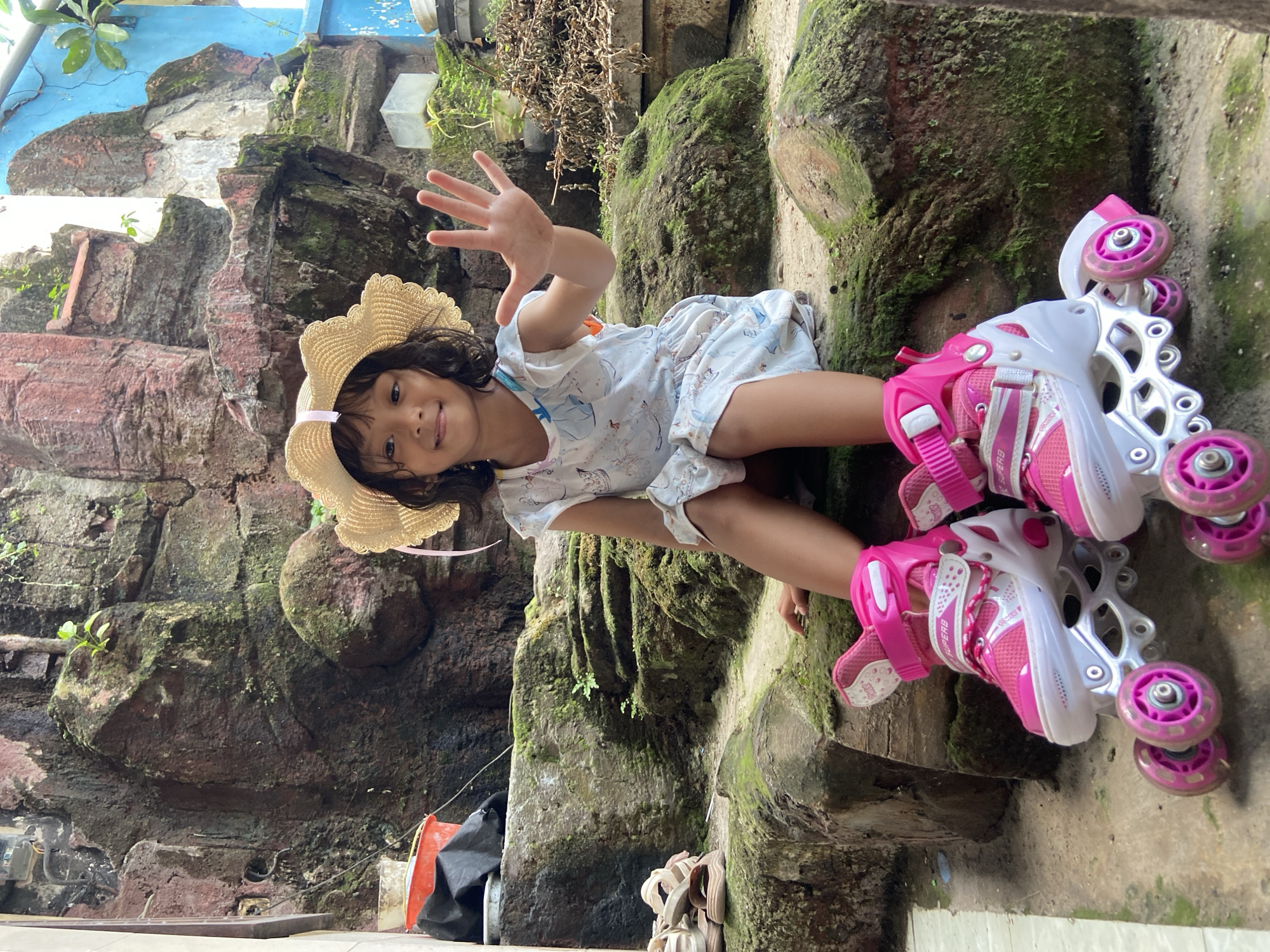 A young girl in a straw hat and a light-colored dress is wearing pink and white roller skates. She is waving her hand at the camera. The location appears to be Gelam, Indonesia, based on visual clues of moss-covered rock walls and tropical vegetation. The setting seems to be an outdoor space with constructed rock features. There are cleaning supplies on the floor in the lower-left corner.