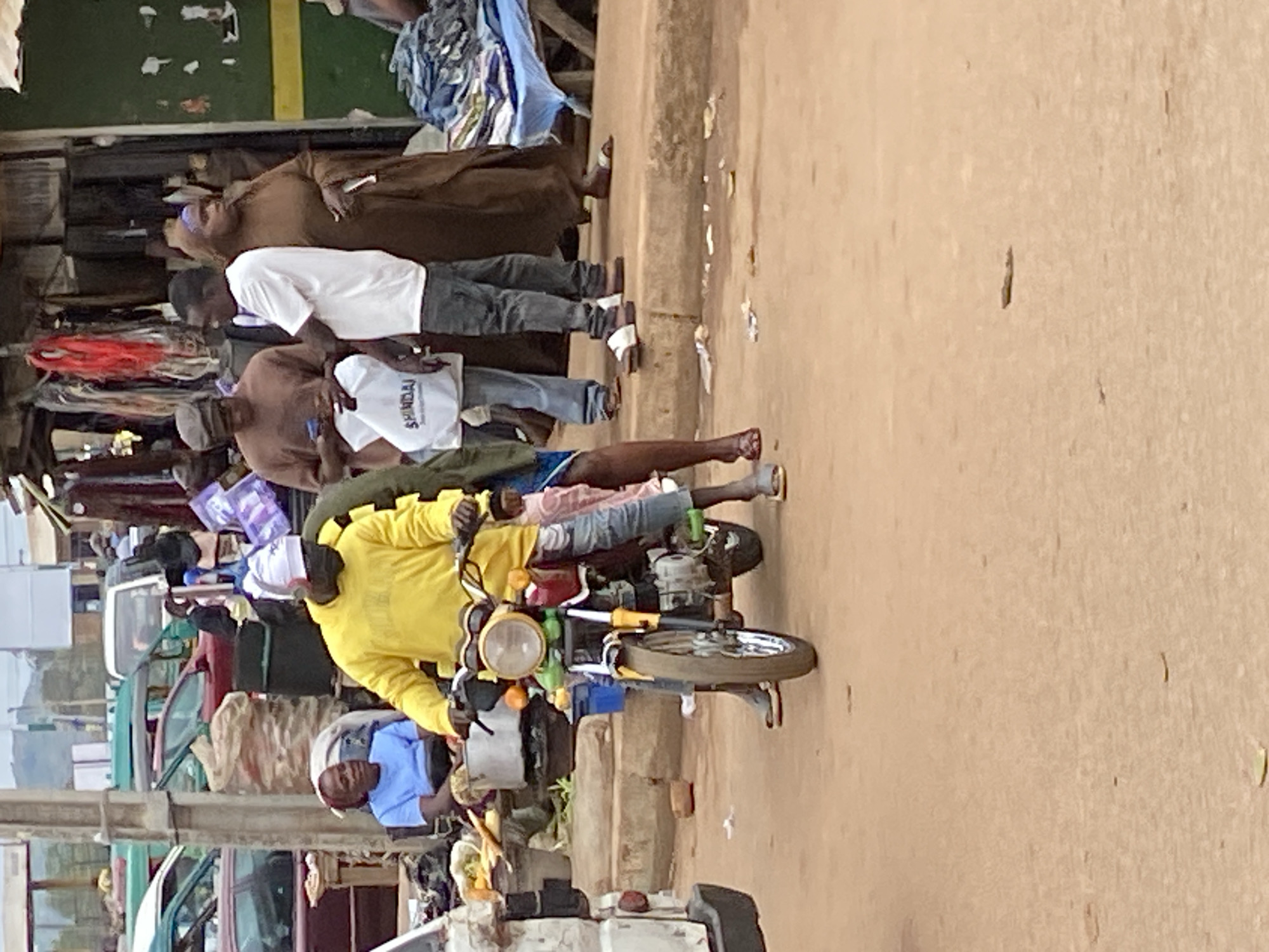 In Bukuru, Nigeria, on a dusty, outdoor market street, a man in a bright yellow long-sleeved shirt and white headwrap rides a red and yellow motorcycle. He is wearing blue jeans and dark sandals, with his hands on the handlebars. The motorcycle has a prominent headlight and orange turn signals. To his right, a woman in a brown garment and headscarf stands holding a white plastic bag. To the left of the motorcycle, a woman in a blue shirt sits on a low stool. Behind them, a stall displays various goods, including fabrics and what appear to be bags of charcoal. Several cars, including a white van and a red car, are parked or driving on the street, and the ground is covered in dirt and scattered debris. The scene suggests a busy market environment with vehicles and pedestrians interacting.