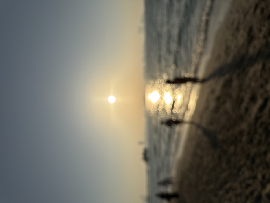 A blurred, wide-angle photograph captures a sunset scene on a beach in Dubai, United Arab Emirates. The bright, circular sun, exhibiting a starburst effect, is centrally positioned just above the horizon. The sky shows a gradient from a light blue-grey at the top to a warm yellow-orange near the horizon line. A large body of water extends from the horizon, with the sun's reflection creating a prominent, shimmering path of bright, circular bokeh highlights on its surface. Gentle waves are visible nearing the sandy shore, which occupies the foreground and appears dark. Multiple heavily blurred human figures are visible on the beach and at the water's edge, appearing as dark silhouettes against the reflective water. Two figures are more prominent in the midground, one positioned to the left of the sun's reflection and another to the right, both casting long, dark shadows across the sand. In the distant background over the water, blurred silhouettes consistent with large vessels or distant coastal structures are faintly discernible on the horizon. The scene depicts activity along a coastal area during sunset.