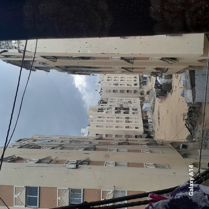 The image depicts a devastated urban landscape in Khan Yunis, Palestinian Territory, viewed from an elevated perspective, possibly a window, with overhead power lines visible. The scene is dominated by multiple multi-story residential buildings under an overcast sky.
In the immediate foreground, a dark structural element partially obscures the right side, and a piece of purple and white patterned fabric is visible on the lower left.
Two prominent multi-story residential buildings frame the central view. The building on the left, beige with light brown trim and decorative white window frames, shows numerous pockmarks on its facade and several damaged or boarded windows. The building on the right, also multi-story, exhibits extensive structural damage, particularly on its upper floors where concrete is shattered and internal elements are exposed. Many of its windows are broken, and some metal shutters are bent.
Between these buildings lies a large, unpaved, sandy or dirt-covered ground area, heavily littered with debris, rubble, and what appears to be collapsed building materials. Within this debris field, several makeshift shelters or tents, constructed from tarpaulins, corrugated metal, and other salvaged materials, are present. Laundry is observed hanging near some of these temporary structures. Further multi-story buildings of similar architectural style are visible in the background, also displaying signs of wear or damage. No individuals are visible in the scene.
