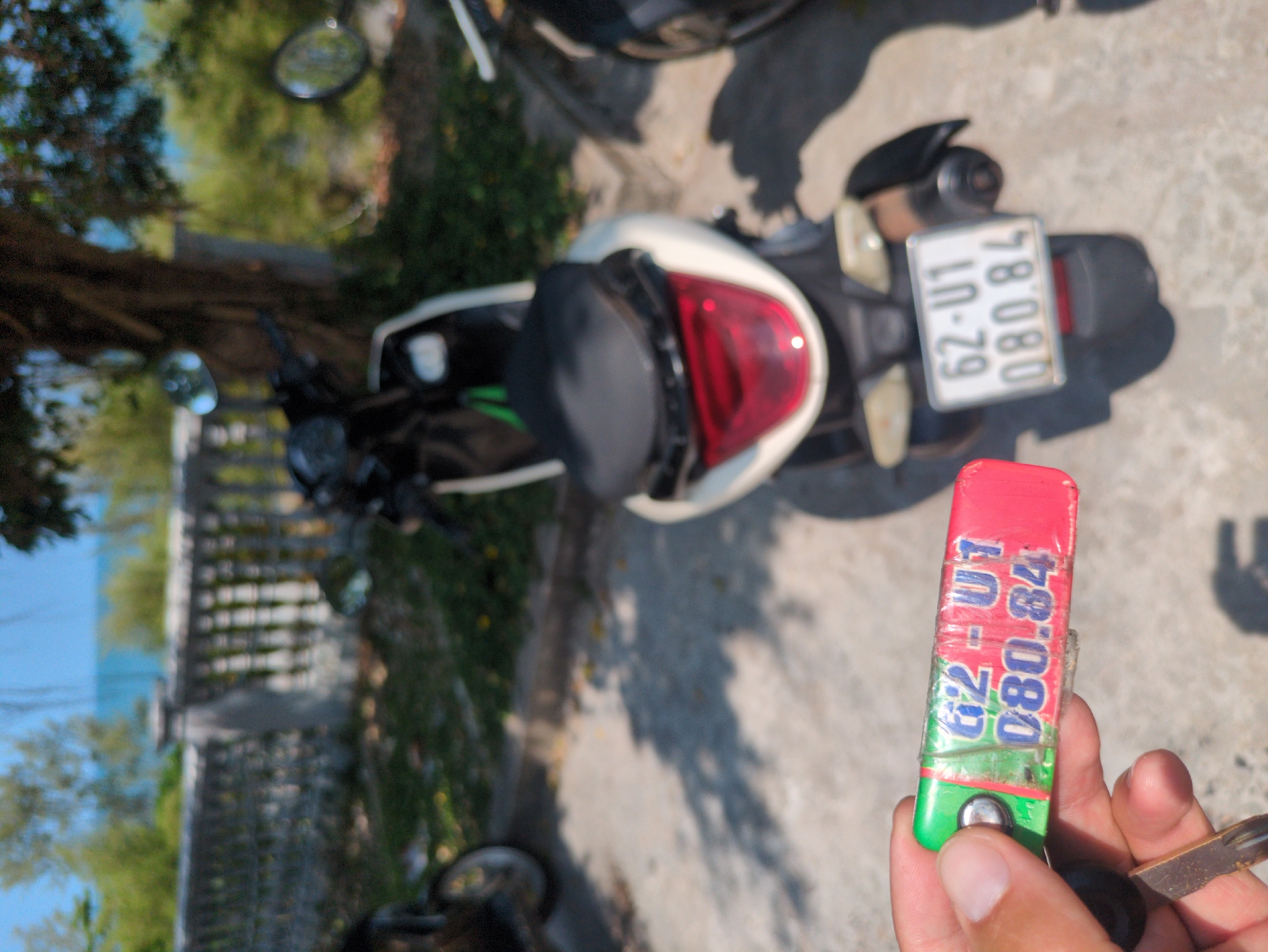 The image shows a person holding a key with the license plate number "62-U1 080.84" written on it. In the background, there is a moped/scooter with a license plate that also reads "62-U1 080.84." There is a tree and some greenery to the left. The setting appears to be outdoors on a sunny day, near what might be a coastal area, judging by the glimpse of blue visible in the distance. This is in Phu Quoc Vietnam due to the presence of Vietnamese license plates, the coastal setting, and tropical plants.