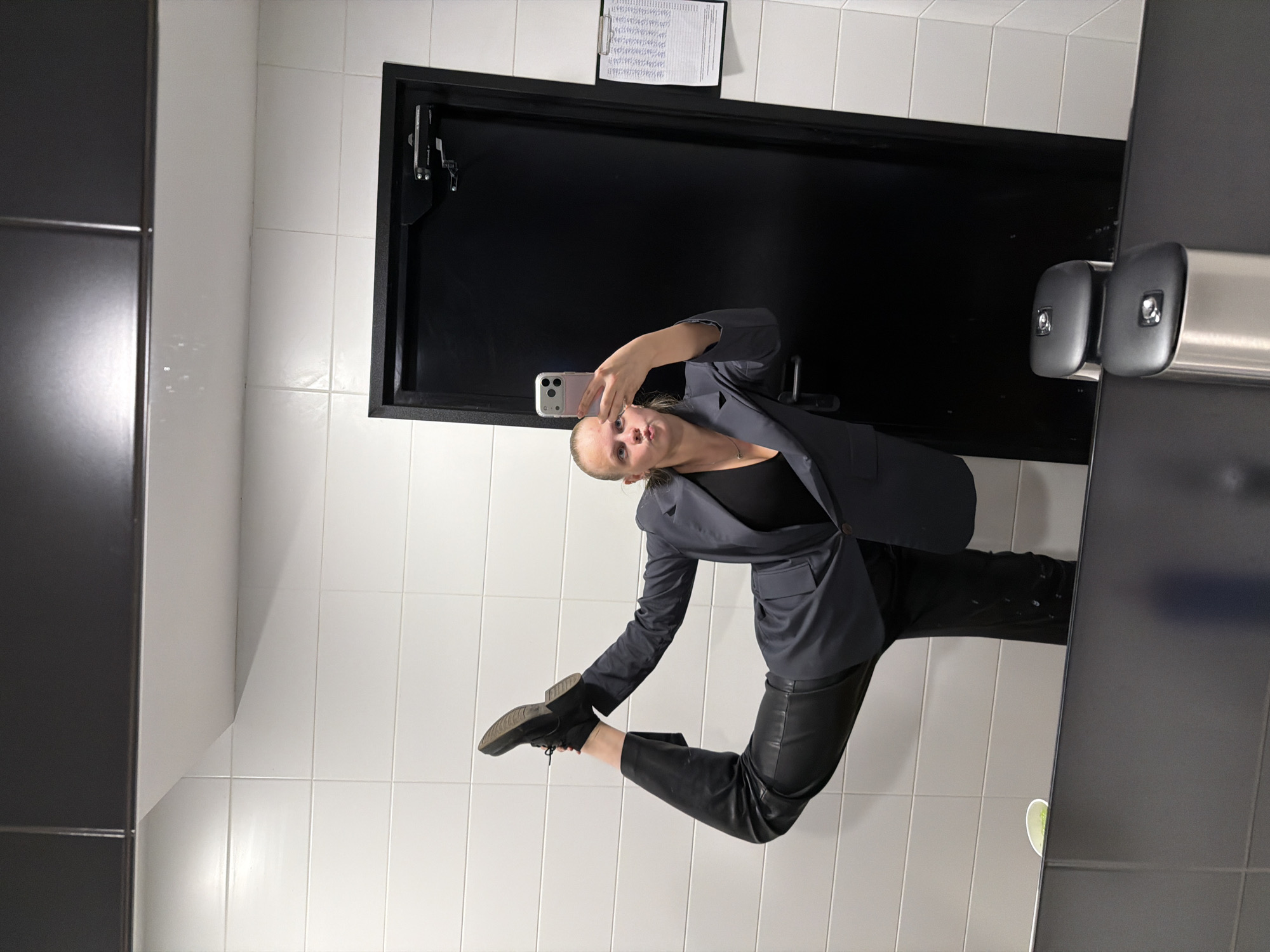 A woman in a gray suit and black leather pants is posing in a bathroom, taking a selfie in the mirror. The bathroom has white tiled walls and a black door with a document hanging on it. The mirror is framed in black and has a metal fixture with two cylindrical objects attached to it. The photo may have been taken in Izhevsk, Russia.