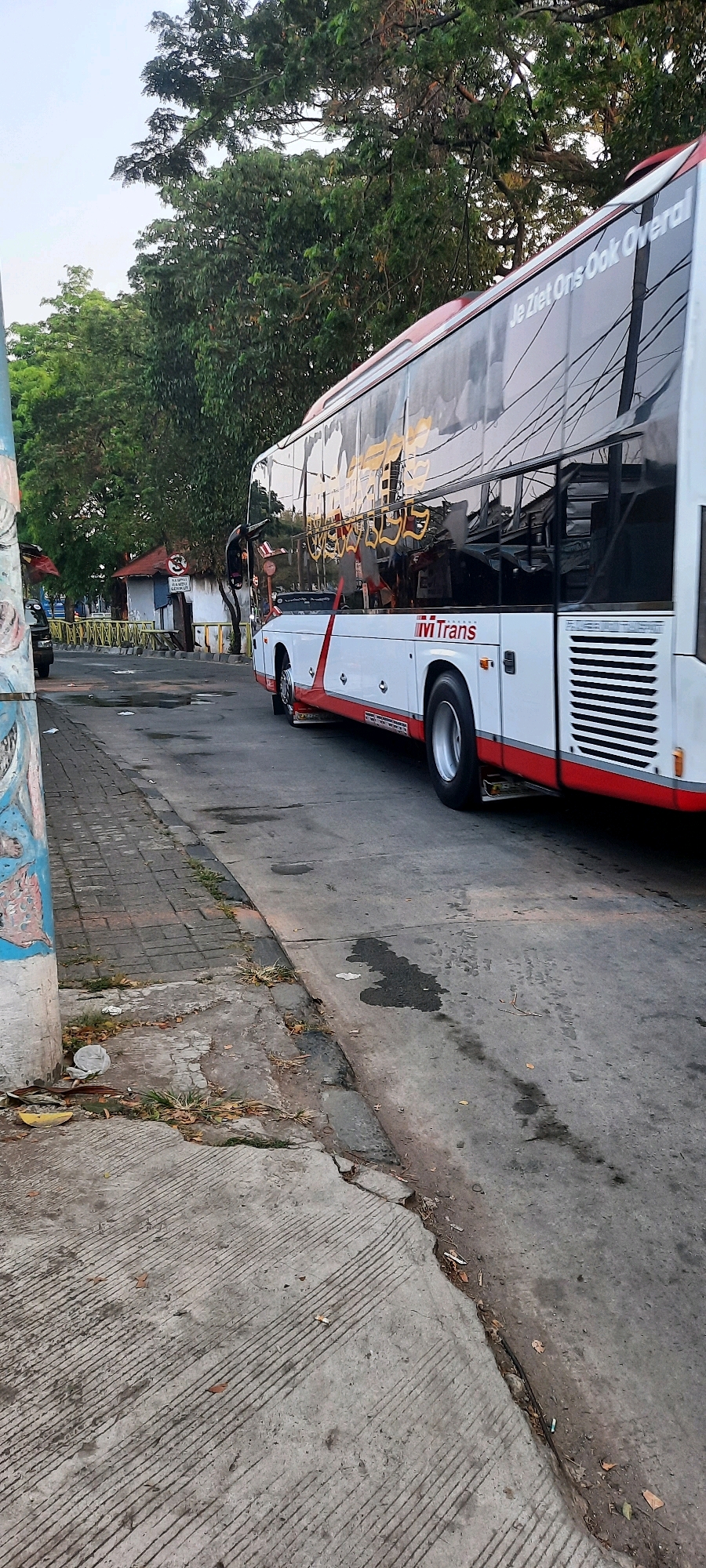 The image shows a street scene in East Jakarta, Indonesia. A large white and red bus with the "ivi Trans" logo and the text "Je Ziet Ons Ook Overal" on its side is parked on the right side of the frame. The bus has tinted windows that reflect the surrounding environment. In the background, there are trees, a small building with a red roof, and a fence. The foreground consists of a concrete sidewalk and a paved road with some water stains. A light pole with graffiti is visible on the left side of the image. The lighting suggests it is daytime, possibly late afternoon.
