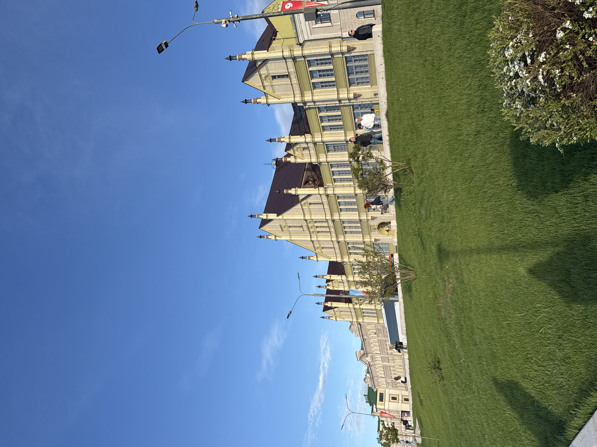 A street scene in Nizhny Novgorod, Russia, shows several matching, light-colored buildings with dark brown roofs. The buildings feature multiple windows and architectural details. A green lawn extends alongside the buildings, with some small trees and a flowering bush. Several people are sitting or standing near the buildings. Streetlights are visible. The sky is a clear blue with some wispy clouds. Flags are displayed near one of the buildings.