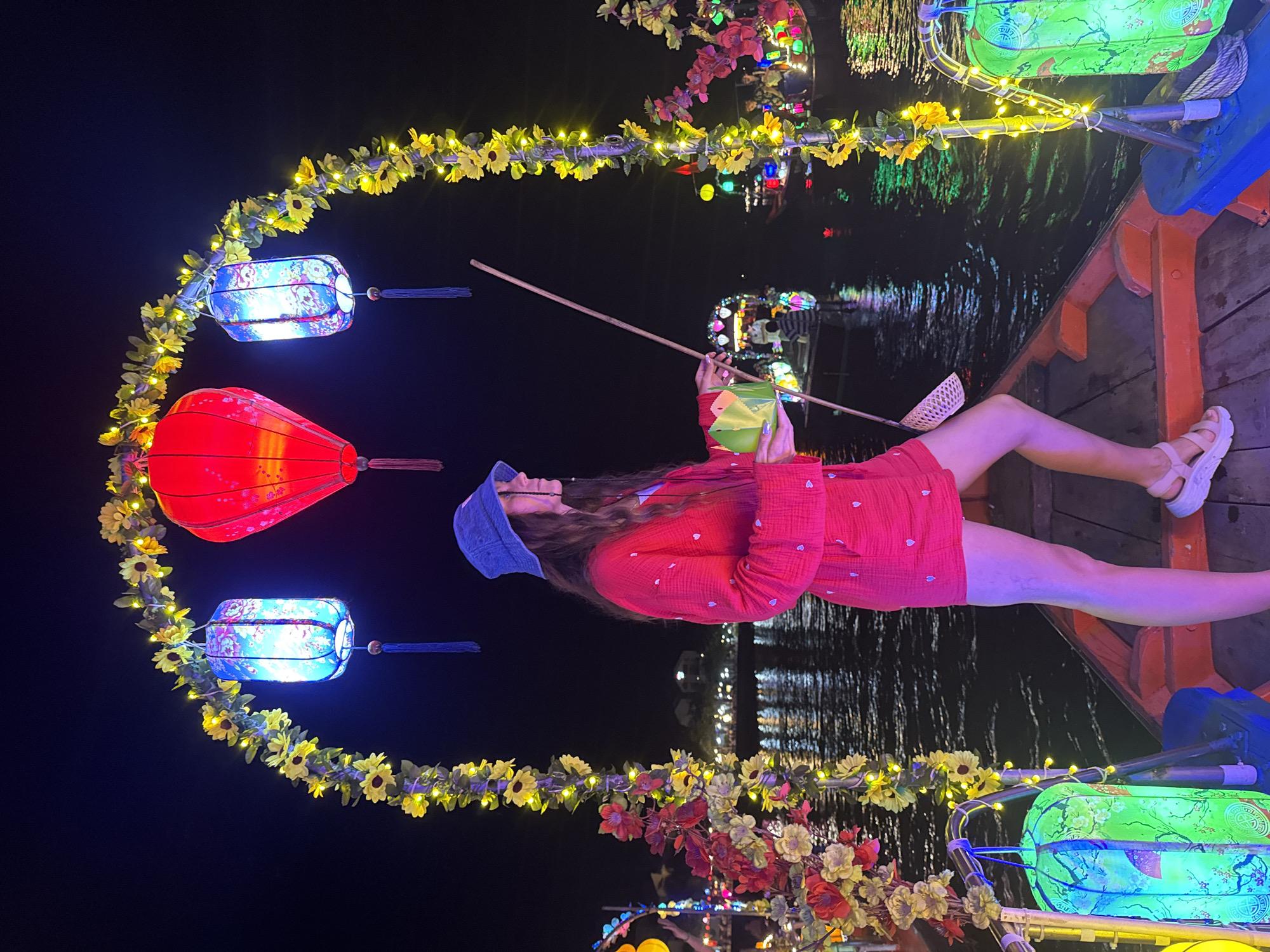 A full-length shot captures a person lying in a boat at night in Hội An Ward, Vietnam. The person is wearing a red dress with white hearts, a blue bonnet, and white sandals, and is holding a long stick with a small net at the end. Lanterns, decorated with yellow flowers and lights, arch over the boat and the water. The river is visible in the background, reflecting the lights of the city.