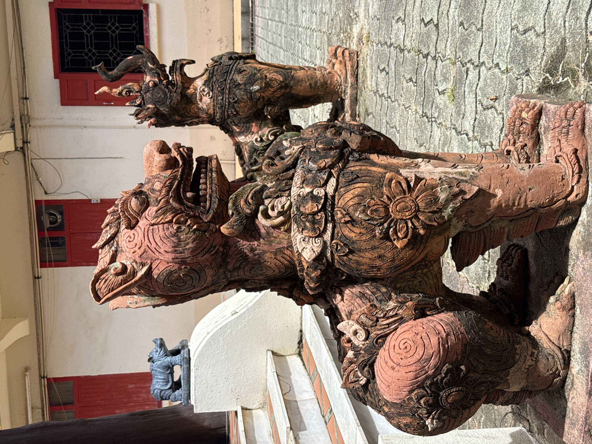Two weathered, terracotta lion-like statues, often referred to as Singha statues in Thai Buddhist tradition, are positioned on a grey, interlocking paver surface in front of a building. The statue in the foreground is larger and more detailed, featuring intricate carvings of floral motifs and swirling patterns on its body and legs. Its mouth is open, revealing sharp teeth, and its mane is elaborately sculpted. The statue in the background is partially obscured and appears to be similar in style but less detailed, with a raised trunk-like appendage on its head.
To the left of the statues, a set of white-painted stairs with red tiled risers leads up to a white-painted platform. Behind the statues, the building features a cream-colored facade with two red-framed windows, one with decorative black grilles and the other with red shutters. A small, dark blue rhinoceros figurine is placed on the white platform to the left, behind the foreground Singha statue. The scene is illuminated by bright sunlight, casting shadows on the ground and highlighting the textures of the statues and paving. This scene is consistent with the grounds of a Buddhist temple or shrine, such as Wat Chalong, located in the Chalong subdistrict of Phuket, Thailand, where such guardian statues are commonly found. There are no people or animals visible in the immediate vicinity of the statues.