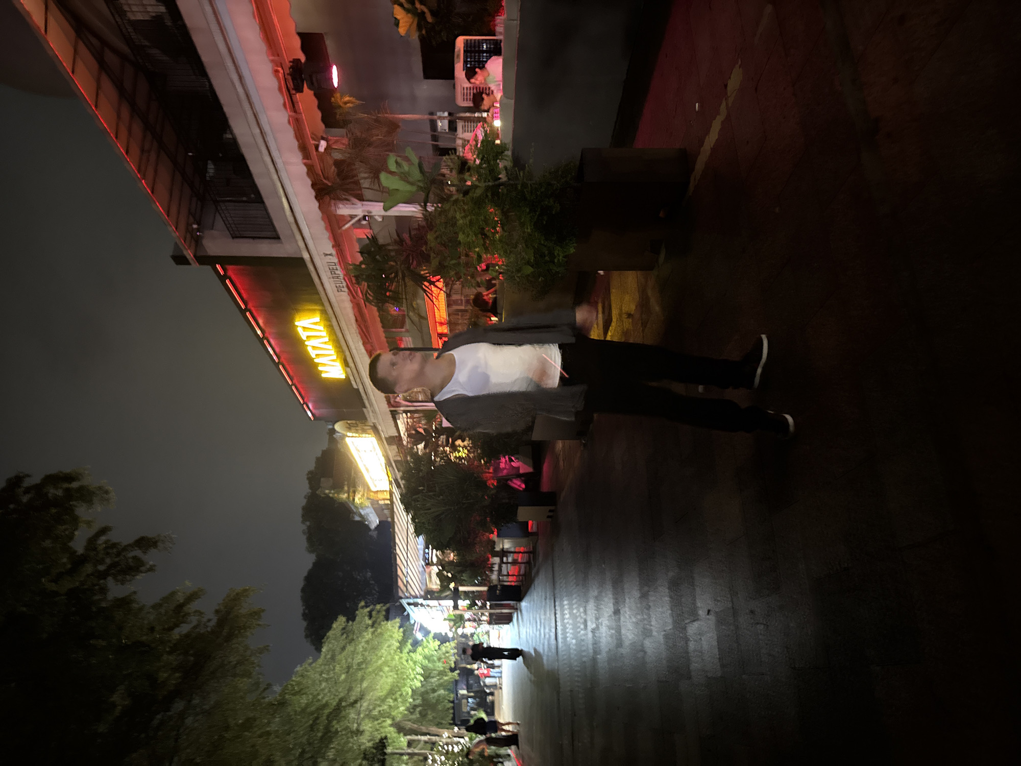 The image, taken at night, shows a street scene in Tianhe District, China. A person in a white shirt, gray jacket, and black pants is standing on a sidewalk. Buildings with illuminated signs are on the left. Trees are on the far left and in the distance. The ground appears to be paved with dark-colored stones. Other people are visible in the distance.